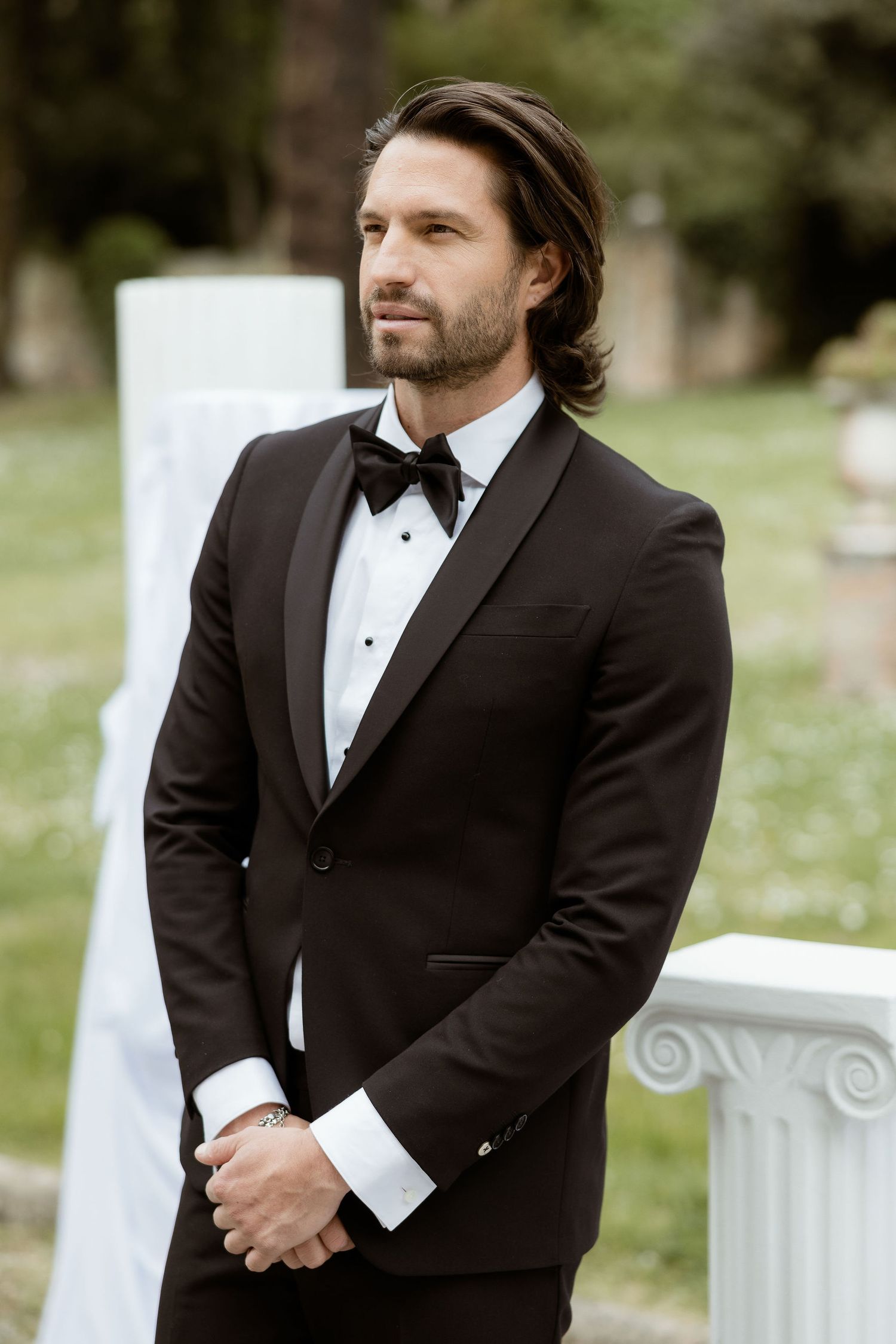 A man in a classic black tuxedo and bow tie poses outdoors with white columns in a garden setting.