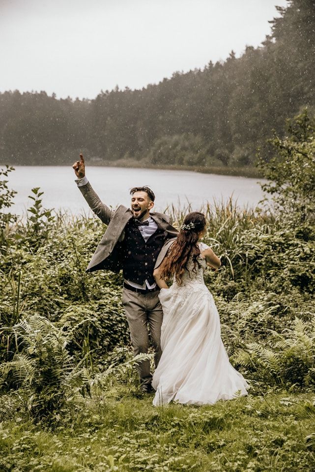 A couple in wedding attire playfully dances and spins together on a grassy hillside overlooking a misty lake.