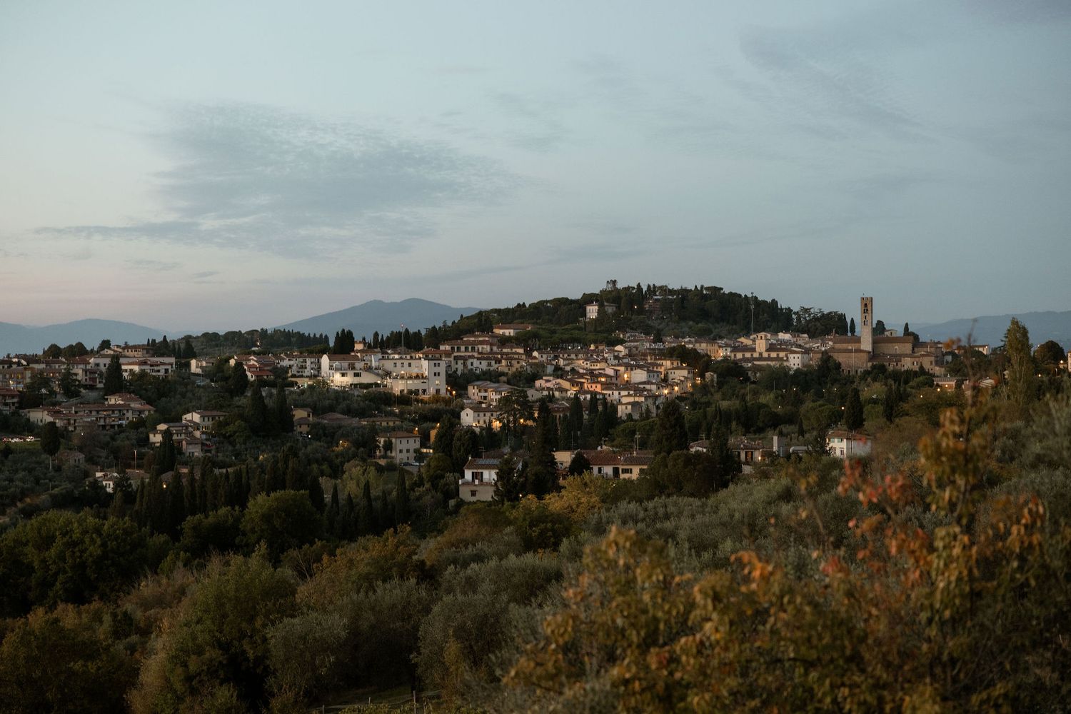 Scenic hilltop view of a Tuscan village surrounded by olive groves at sunset with historic buildings and church spires.