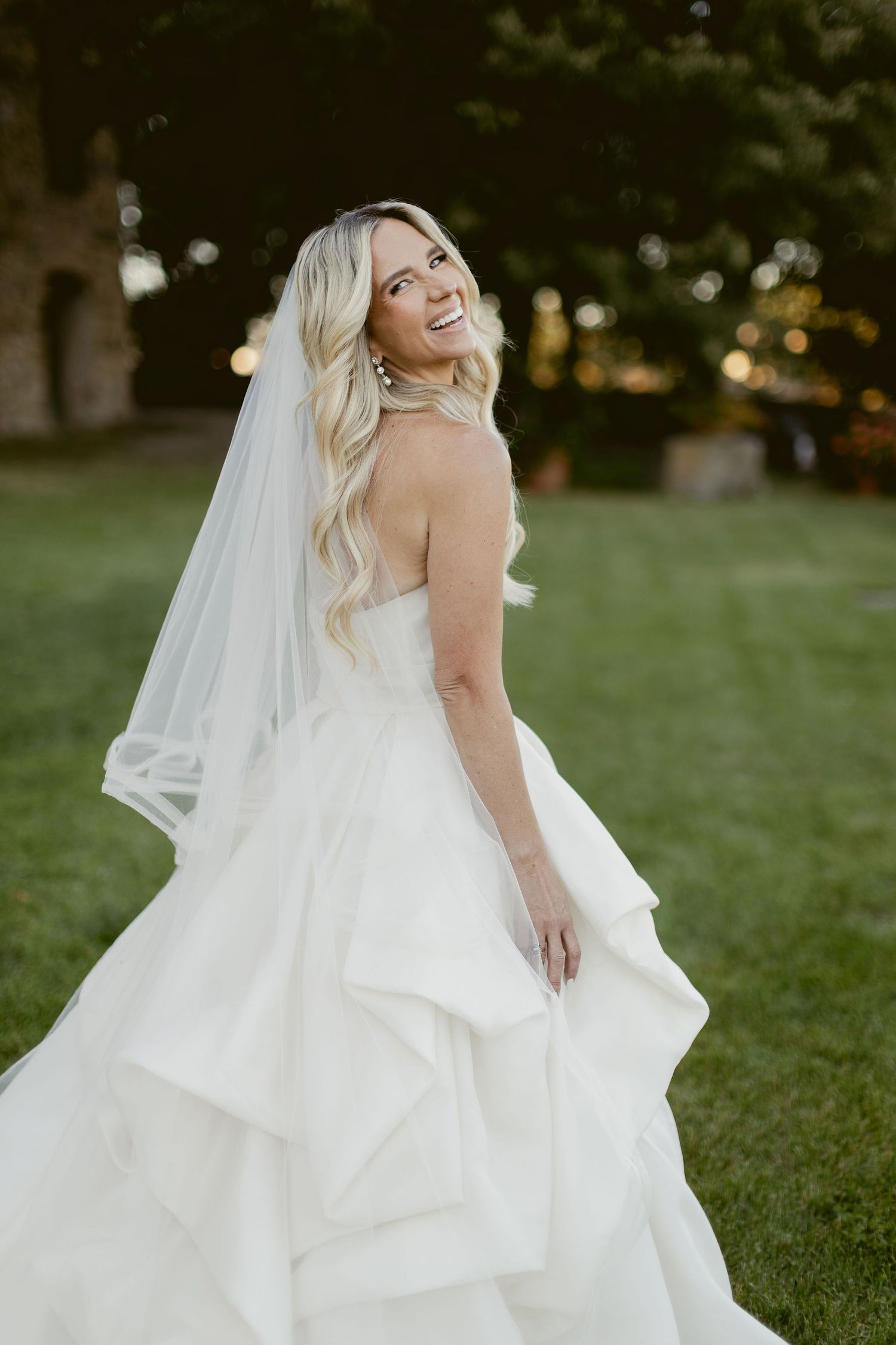 A bride in a flowing white wedding gown with ruffled layers twirls joyfully on a grassy lawn at sunset.