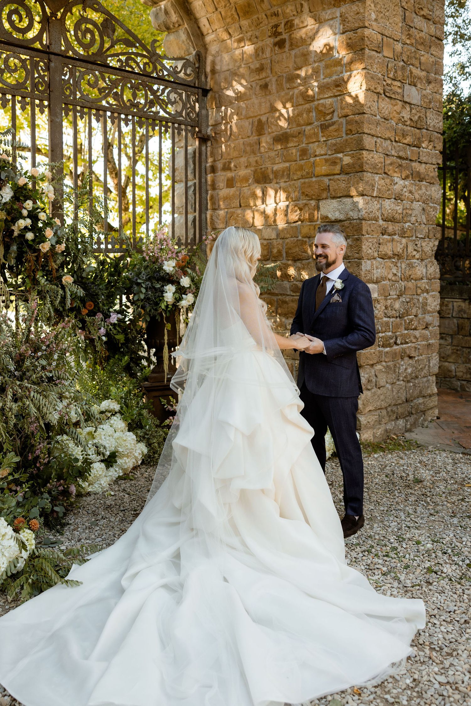 A romantic wedding couple shares an intimate moment in a stone archway garden setting surrounded by white floral decor.