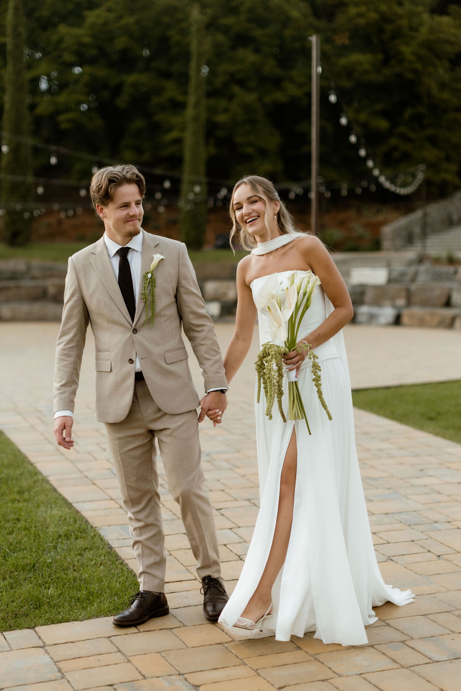 A couple in wedding attire holds hands while walking on stone pavers outdoors during their special day.