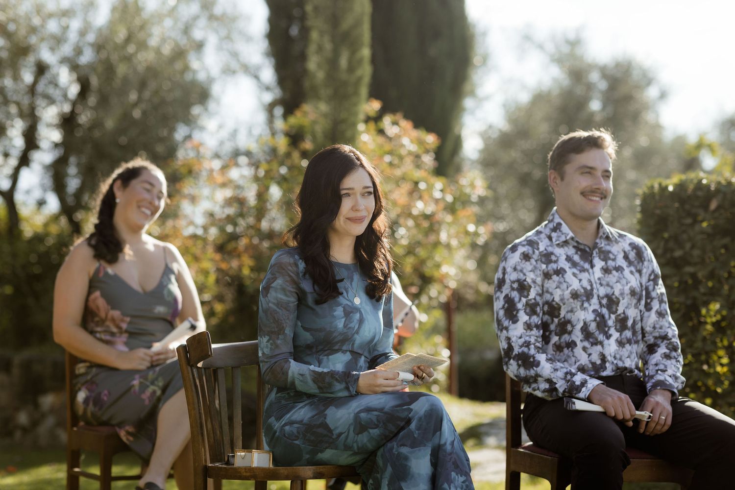 Three people sitting on wooden benches in a sunny garden with lush greenery and trees in the background.