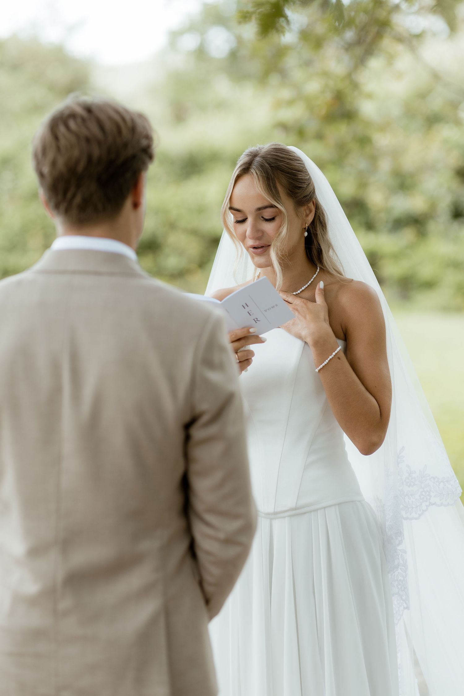 Emotional first look moment captured between bride in white dress and groom in beige suit during outdoor wedding ceremony.