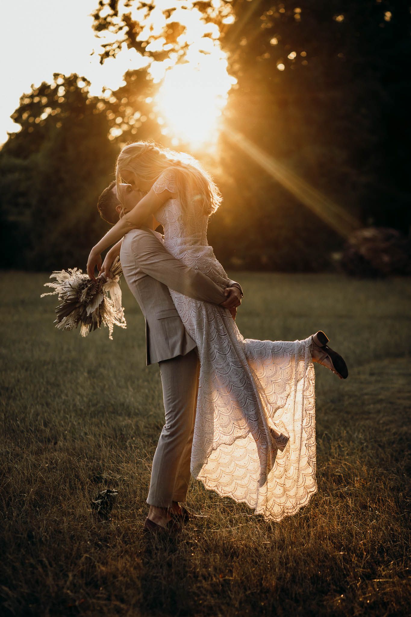 A couple embraces in a romantic sunset moment, with a lace wedding dress flowing in the golden evening light.