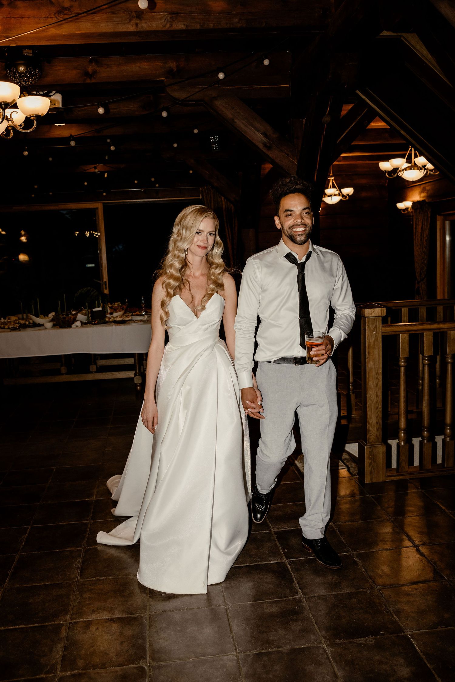 Wedding couple walks together on dark hardwood floors in rustic venue with exposed beams and warm lighting overhead.