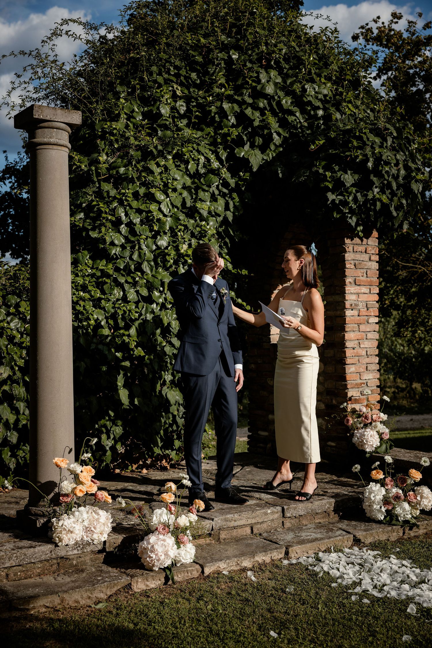 A couple exchanges vows under a garden archway decorated with small figurines.