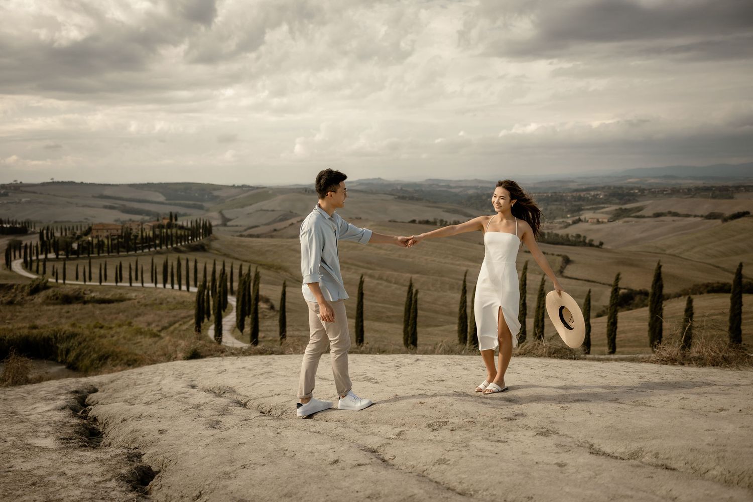 A romantic couple in light summer outfits walking along a scenic Tuscan countryside path with cypress trees.