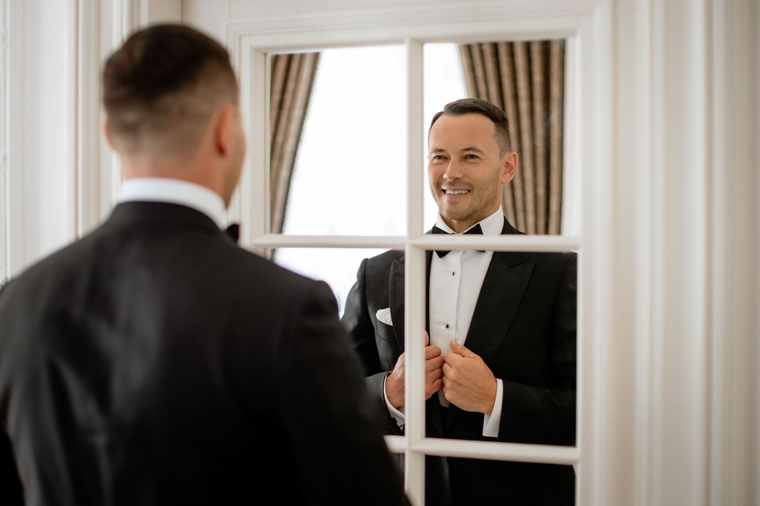 Groom in tuxedo adjusts bowtie while looking in mirror during wedding preparation.