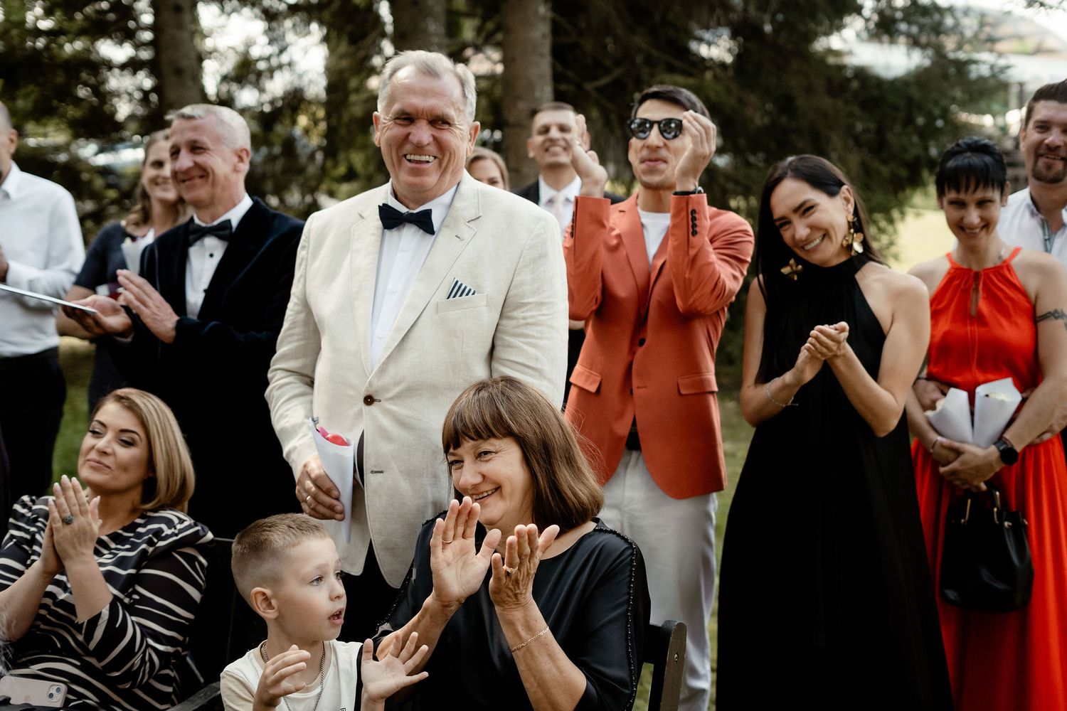 Wedding guests clap and celebrate during an outdoor ceremony with tall trees in the background.