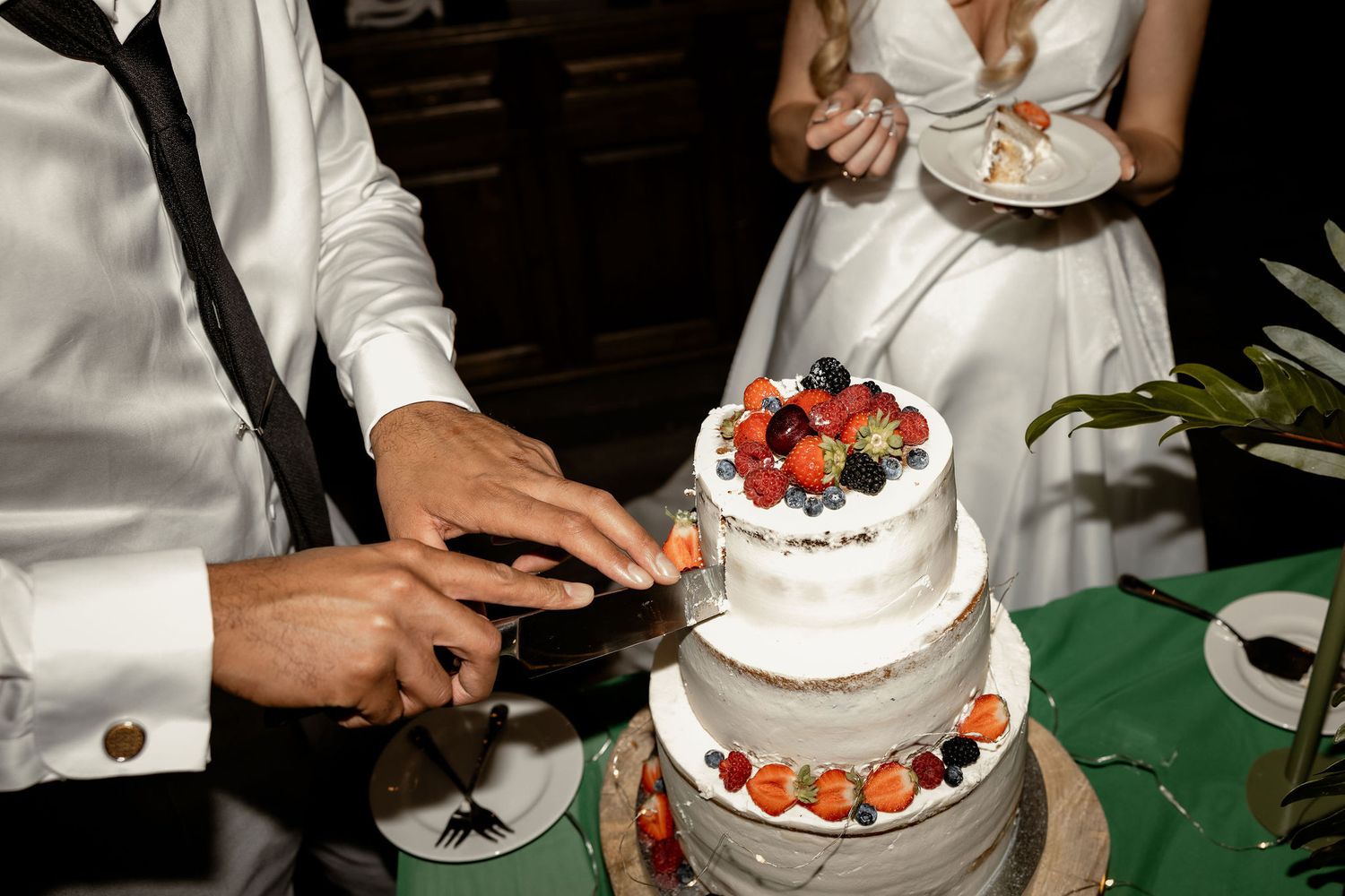A wedding couple cuts into their three-tiered white cake decorated with fresh strawberries on top.