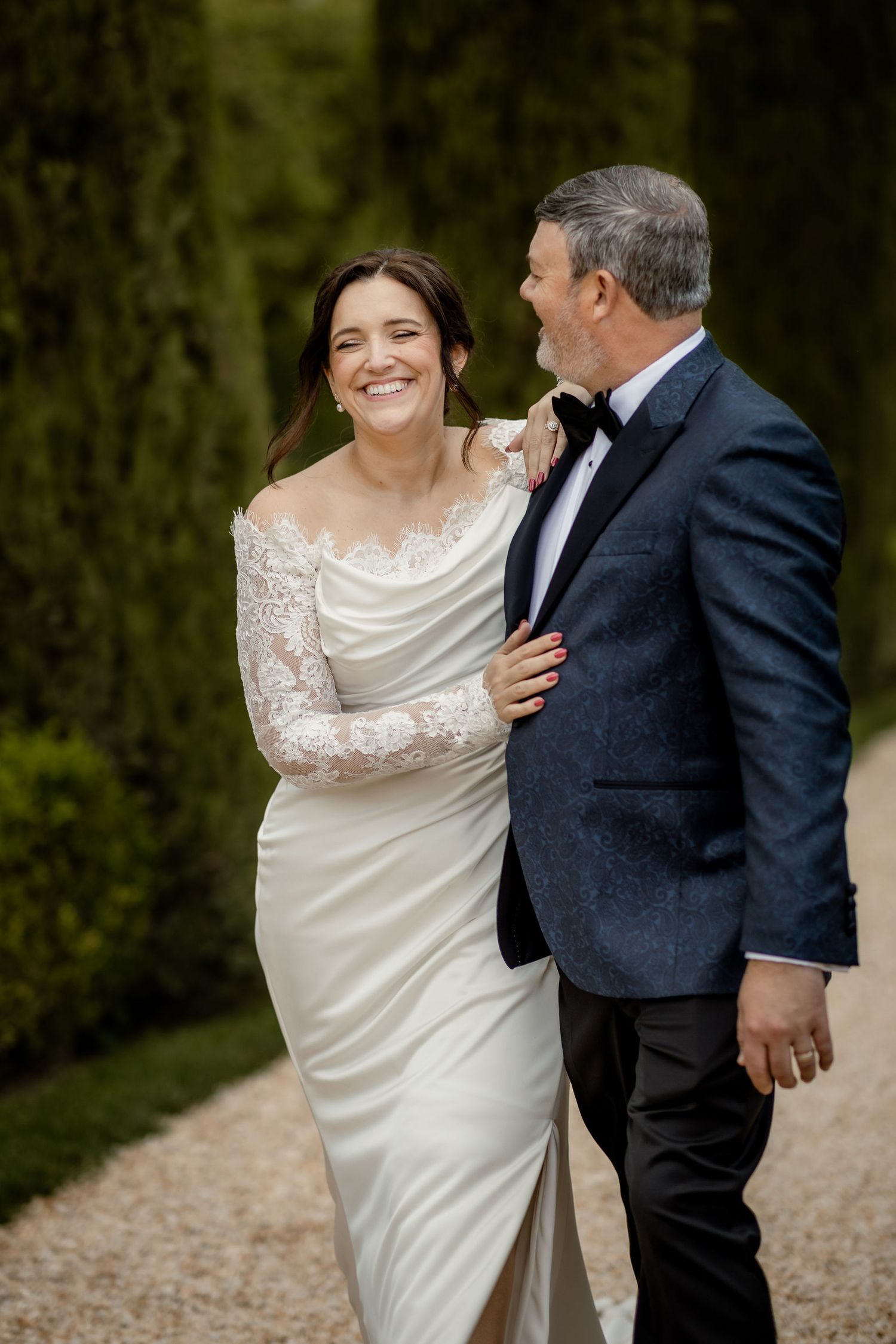 A bride in a white lace-sleeved gown and groom in a tuxedo share a joyful moment on a gravel path.