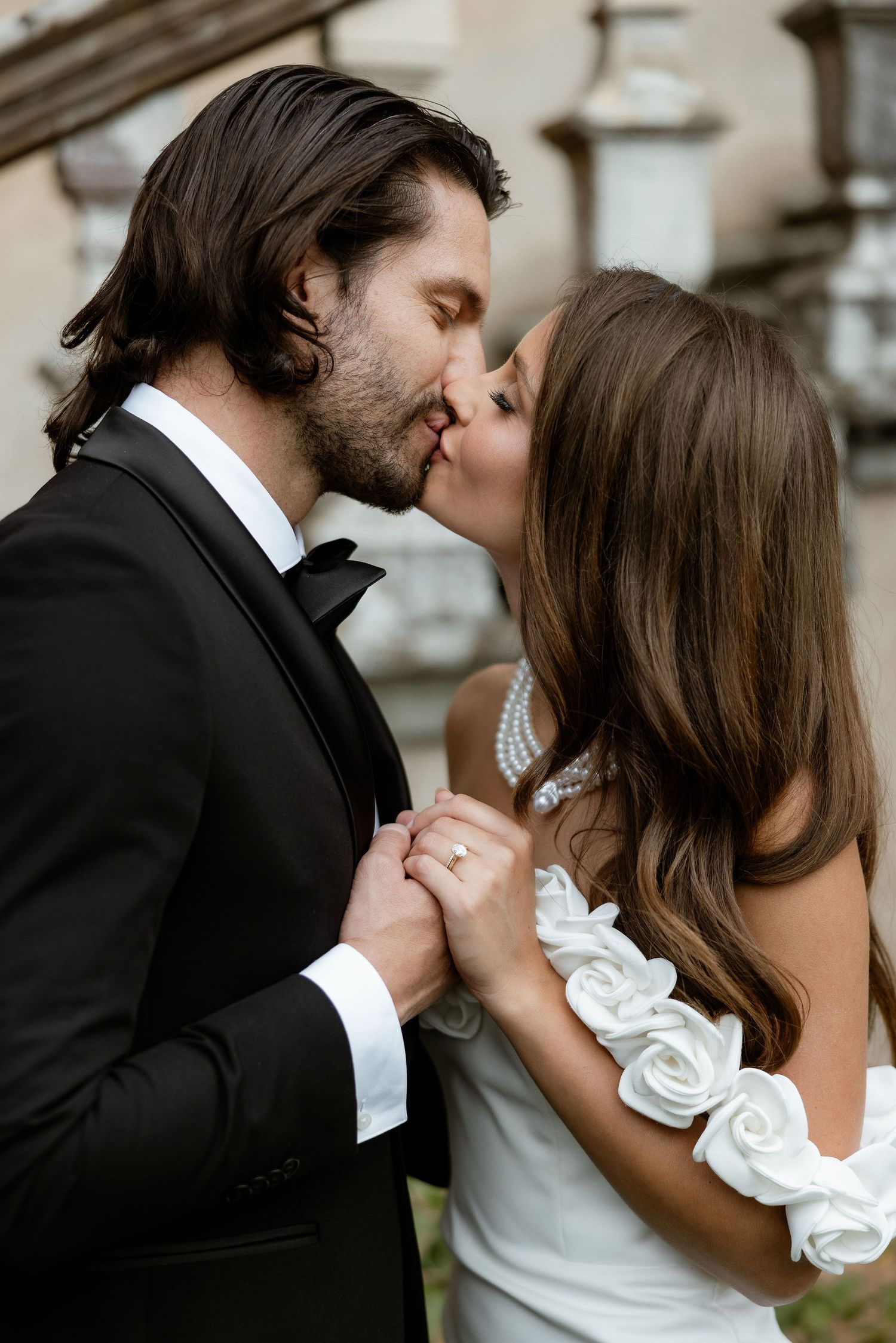 Romantic couple in wedding attire share an intimate kiss while holding hands outdoors with ornate architecture behind them.