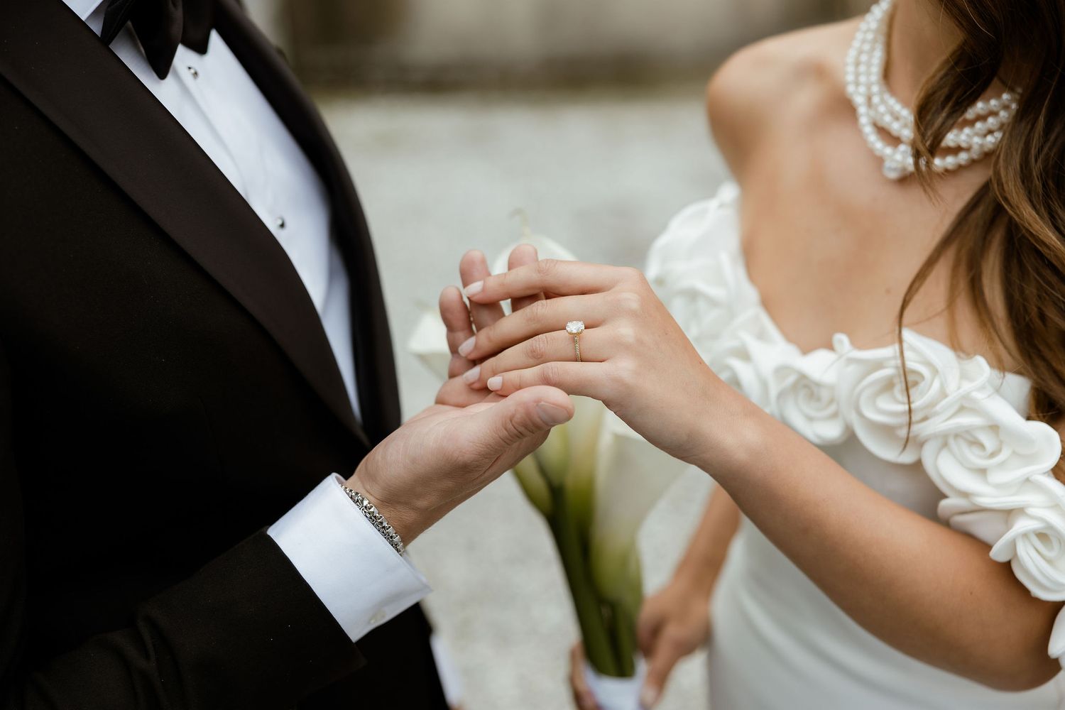 Close-up view of hands exchanging wedding rings during marriage ceremony with white lace dress and black suit details.
