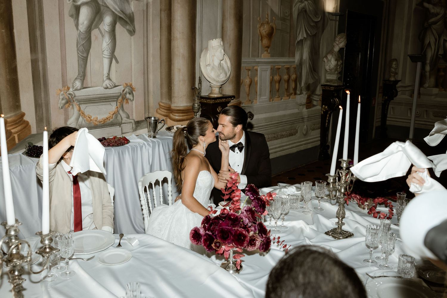A couple in formal wedding attire shares an intimate moment at an elegant dinner table with white linens and candelabras.