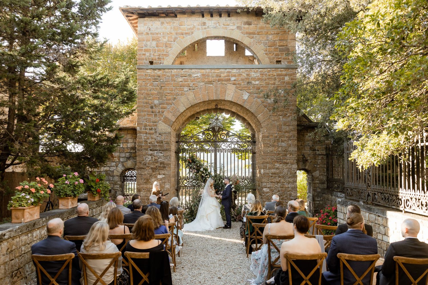 A rustic outdoor wedding ceremony setup with wooden chairs and stone archway surrounded by lush greenery.