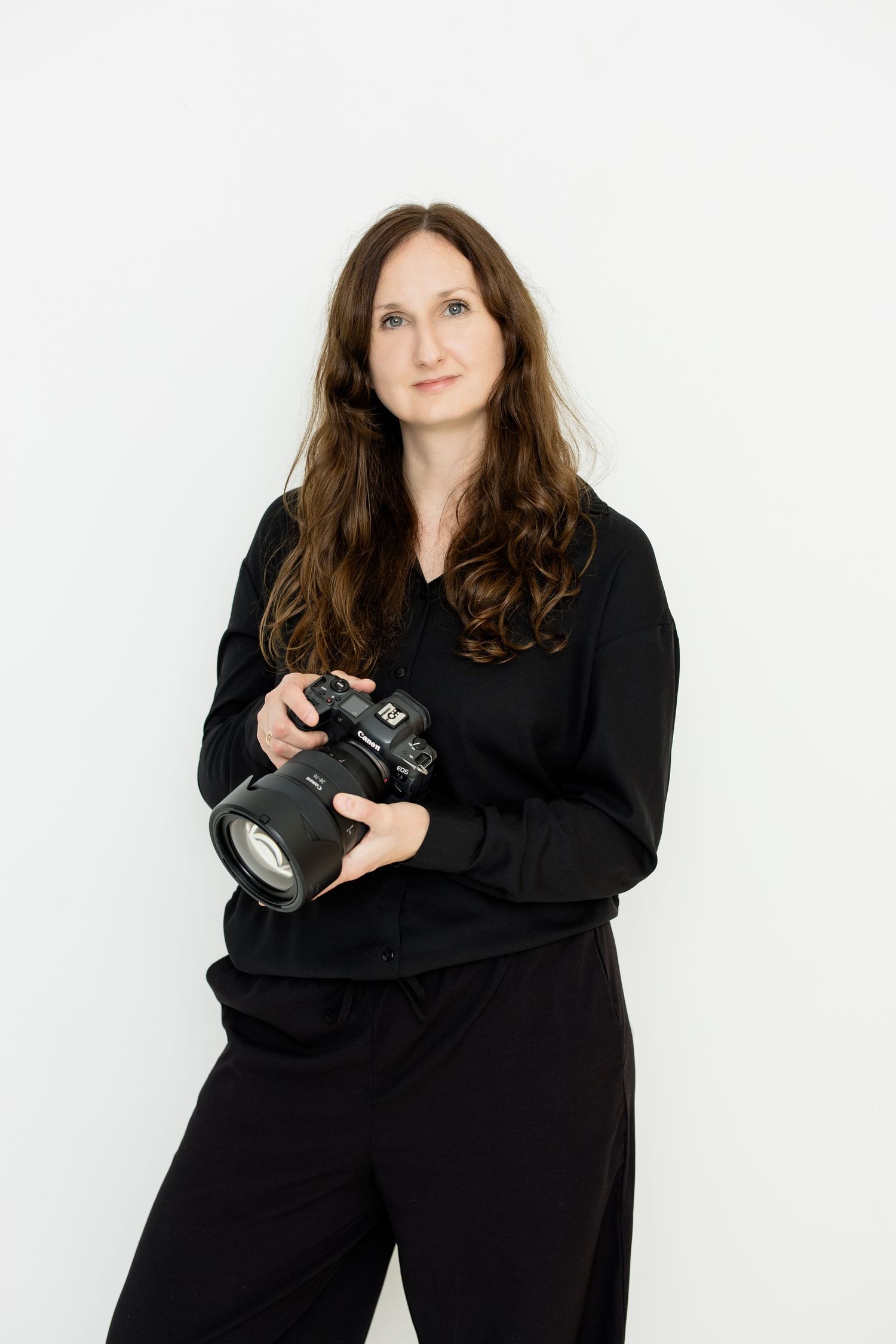 Professional photographer in black attire holds a DSLR camera against white studio backdrop.