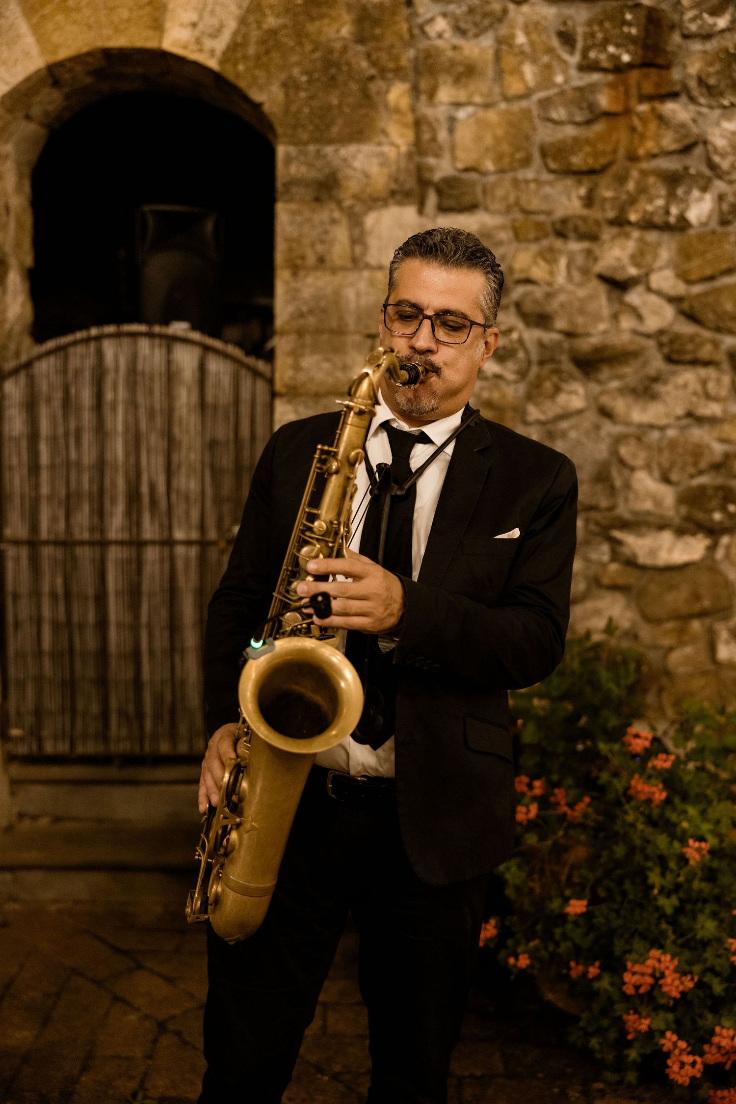 A jazz musician in a black suit plays saxophone against a rustic stone wall with blooming flowers.