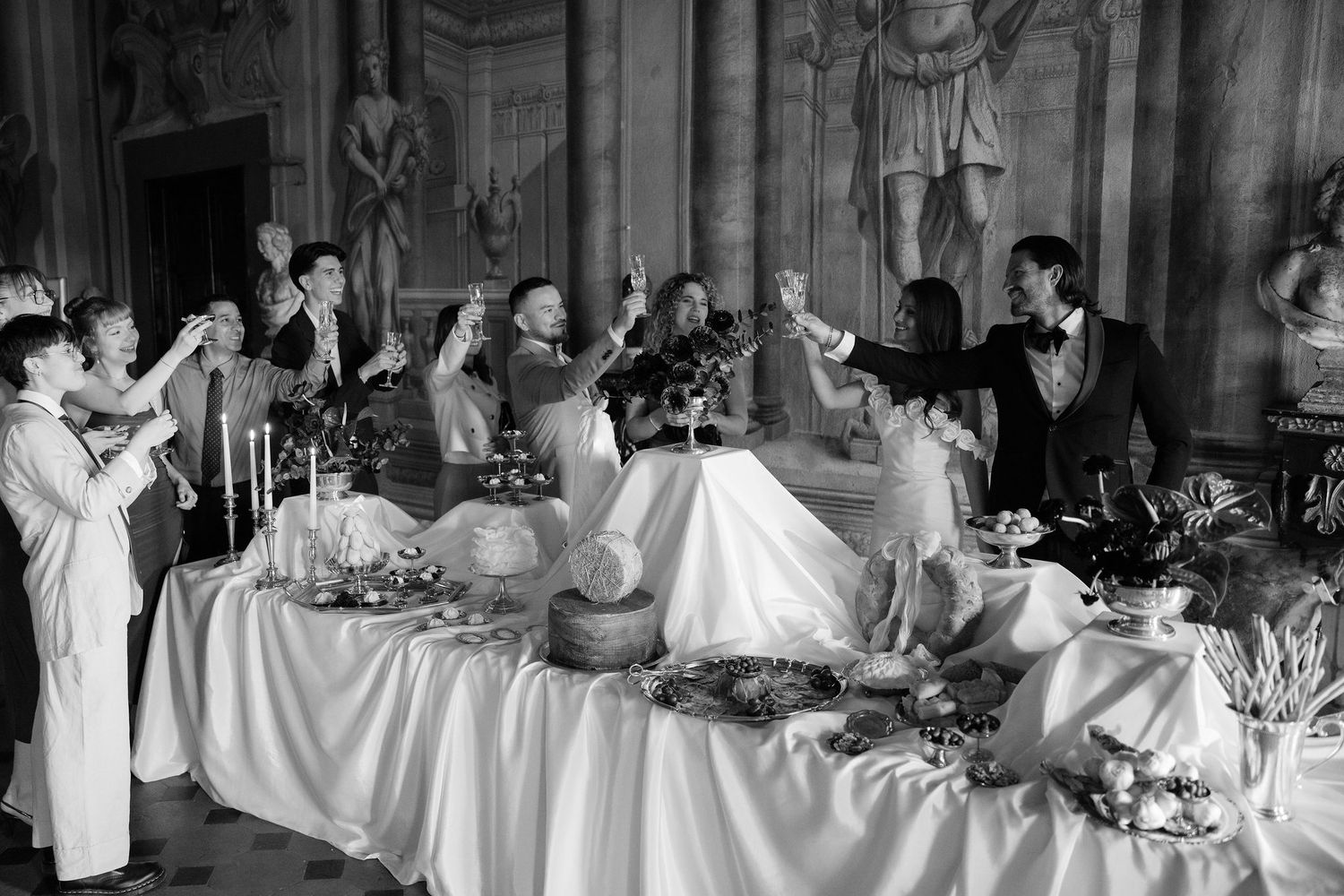 Guests celebrate around an elegant banquet table in a grand room with statues and decorative architecture.
