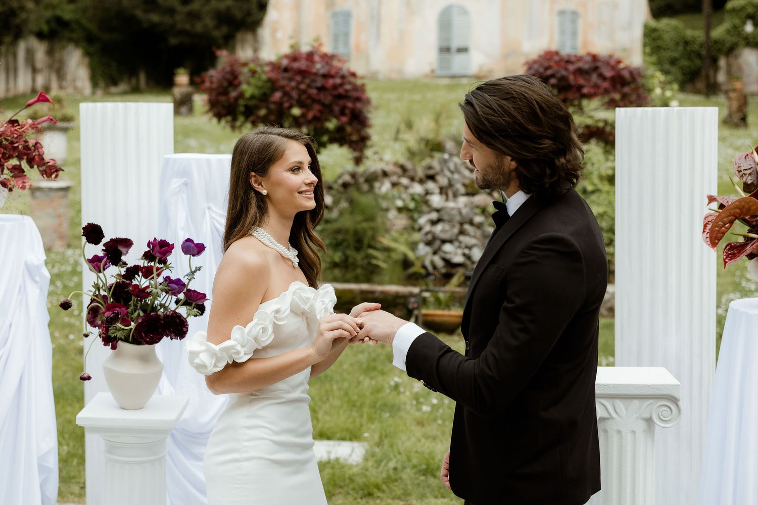 A romantic outdoor wedding ceremony with white columns and deep burgundy floral arrangements in a garden setting.
