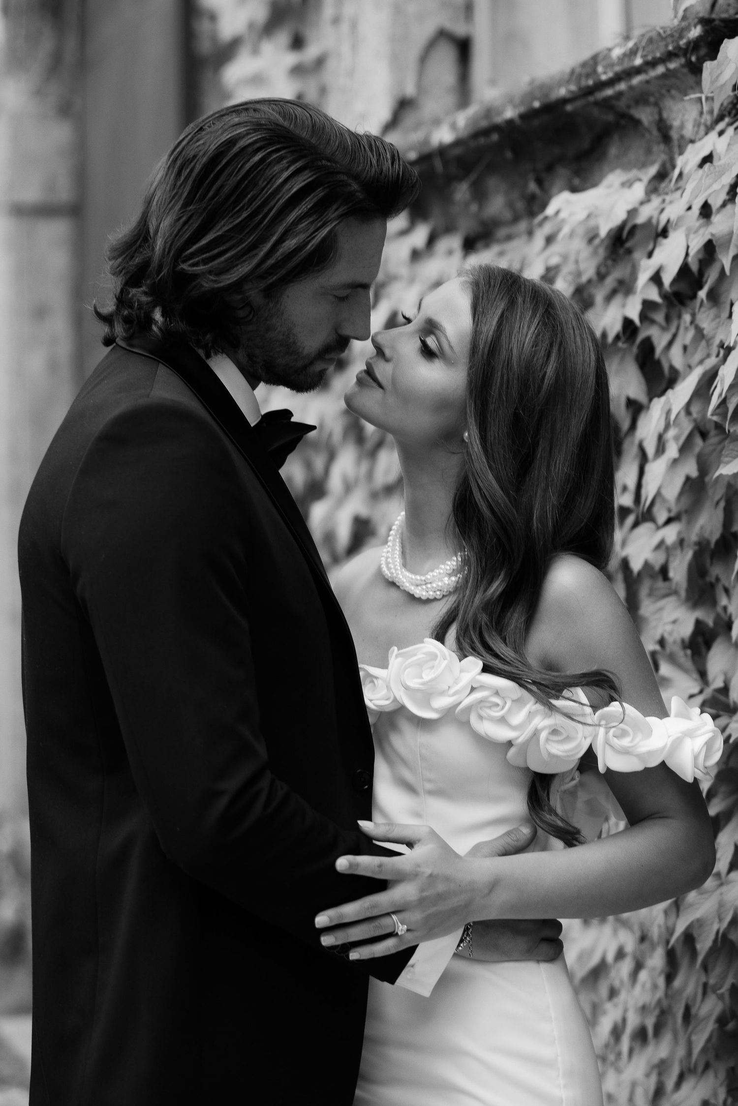 Black and white romantic photo showing a couple sharing an intimate moment by an ivy-covered wall in formal attire.
