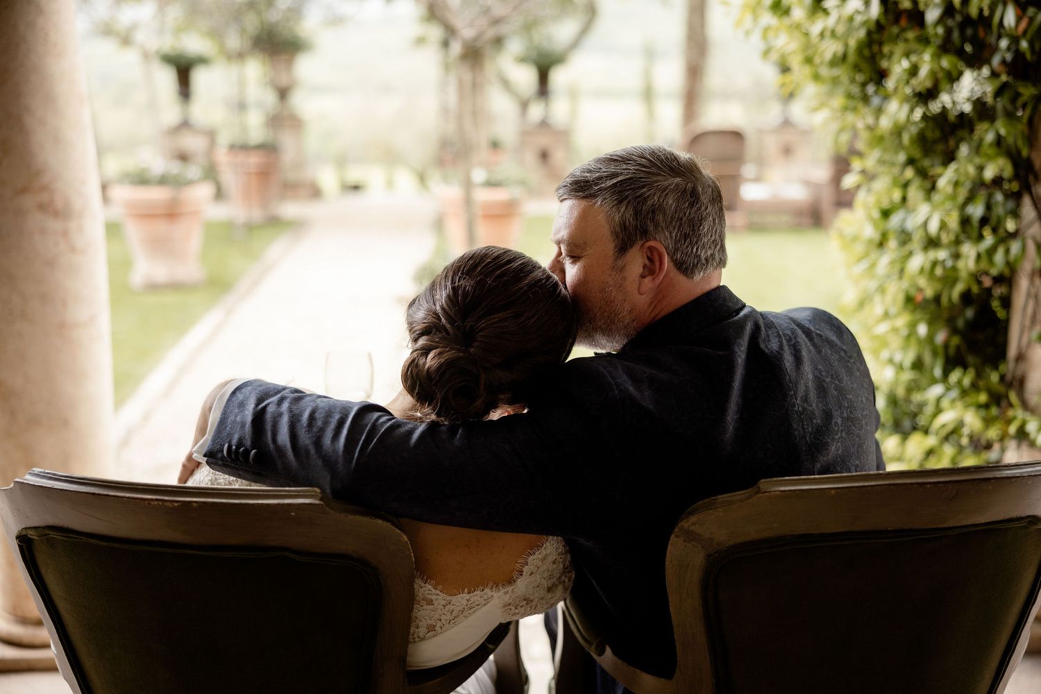 Two people share an intimate moment while sitting together on wooden chairs in a garden setting.