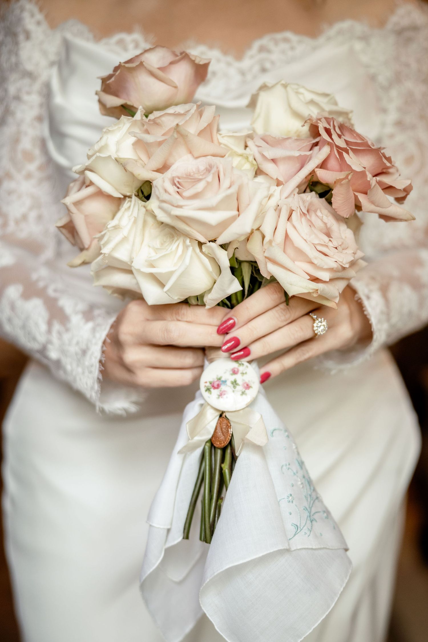 A bride holds a soft blush and cream rose bouquet wrapped in white lace, showing red manicured nails and engagement ring.