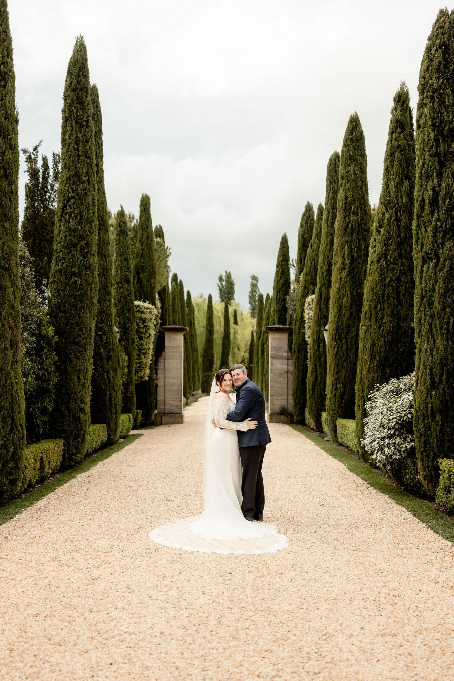 Couple shares romantic moment along cypress tree-lined path at elegant Italian-style villa garden.