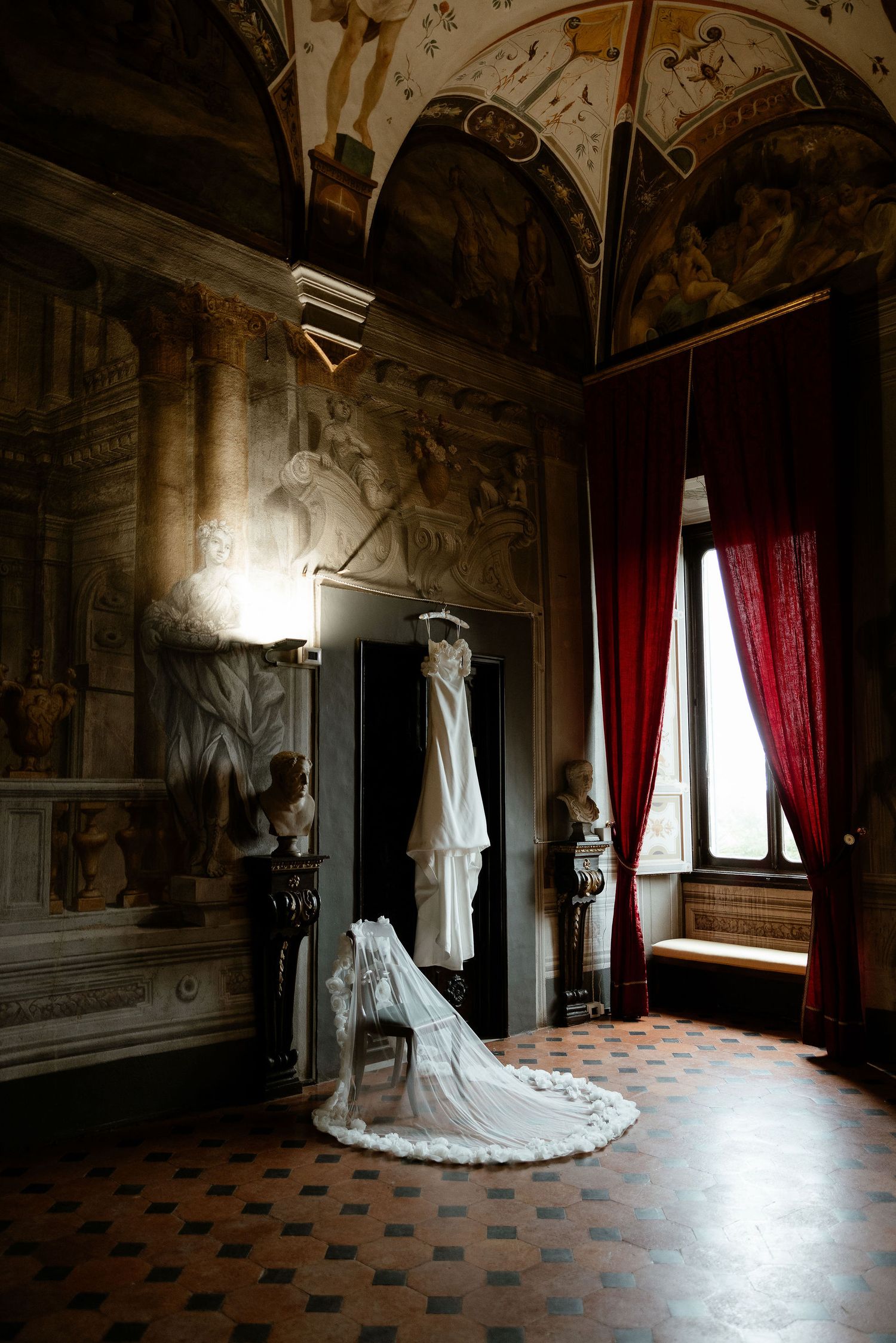 Elegant white wedding dress hanging in a grand historic room with frescoed walls, red curtains and checkered floors.