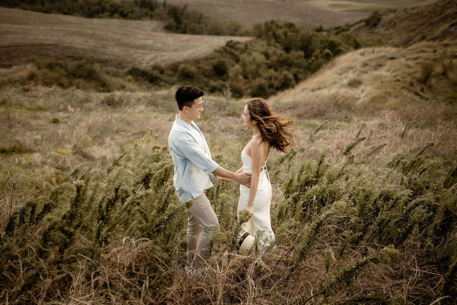 A couple holds hands while walking through a windswept grassland.