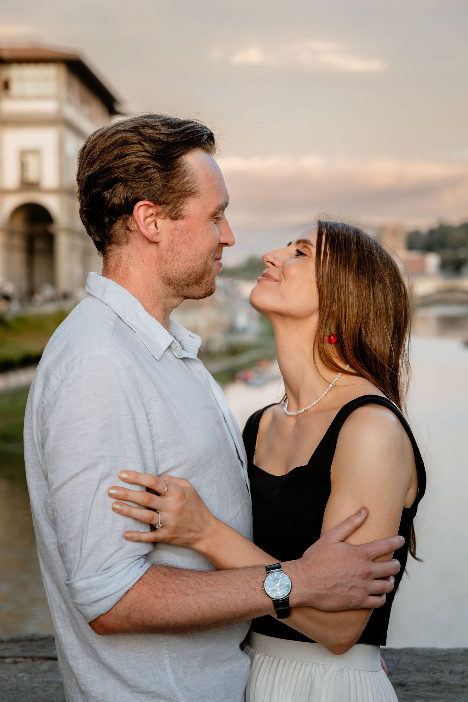 A romantic couple embraces during sunset in Florence, Italy with warm lighting and the Arno river in the background.