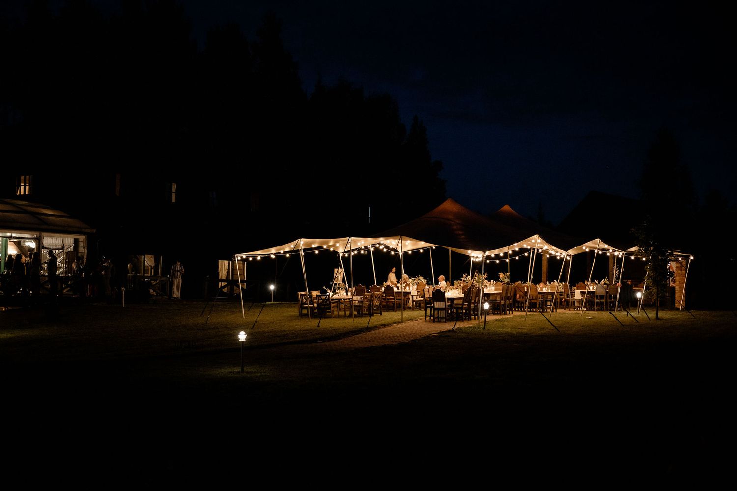 String lights illuminate an outdoor wedding tent and reception area against a dark night sky.