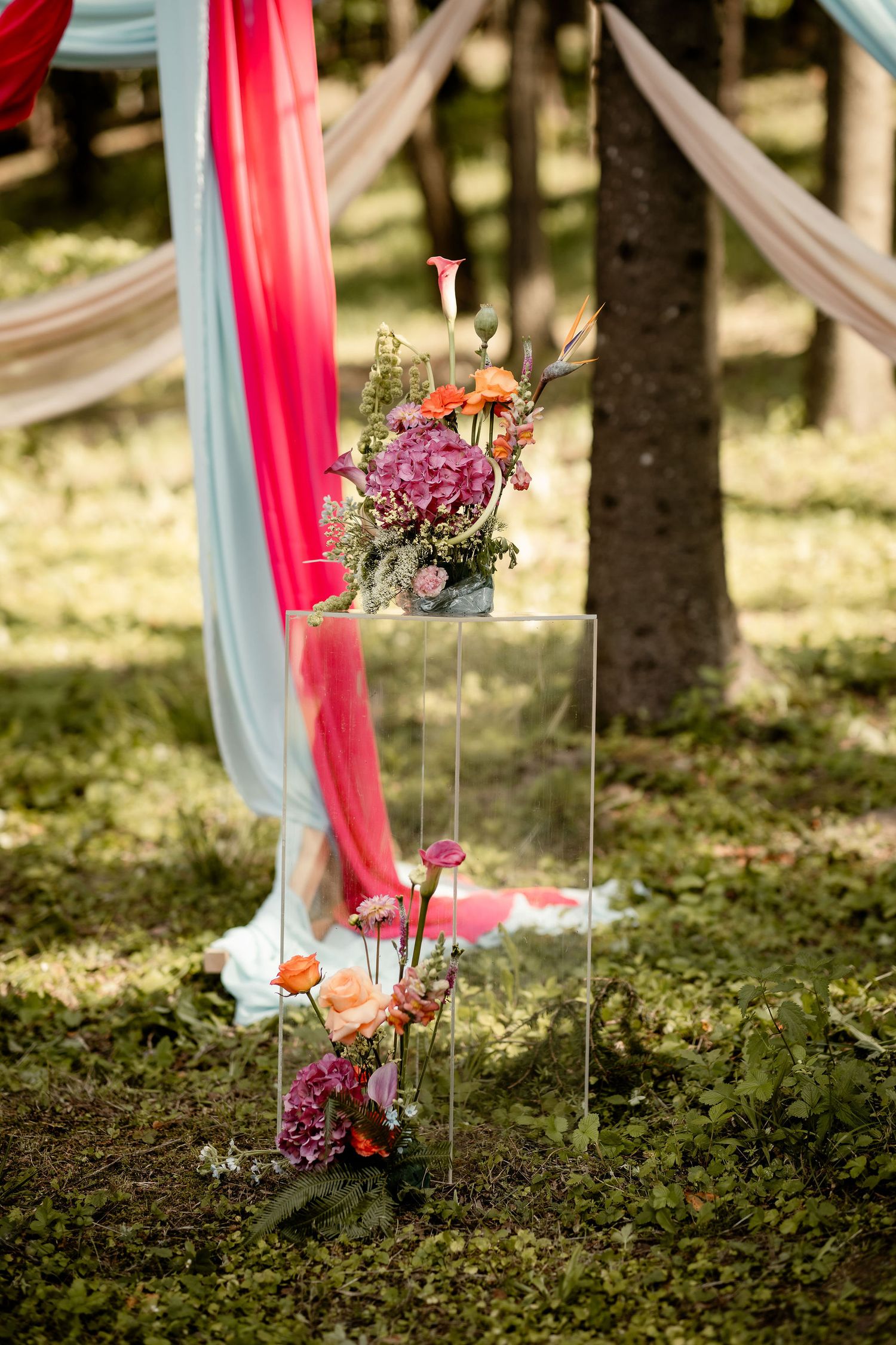 Outdoor wedding decoration with pink flowers on clear acrylic stands and flowing fabric drapes.