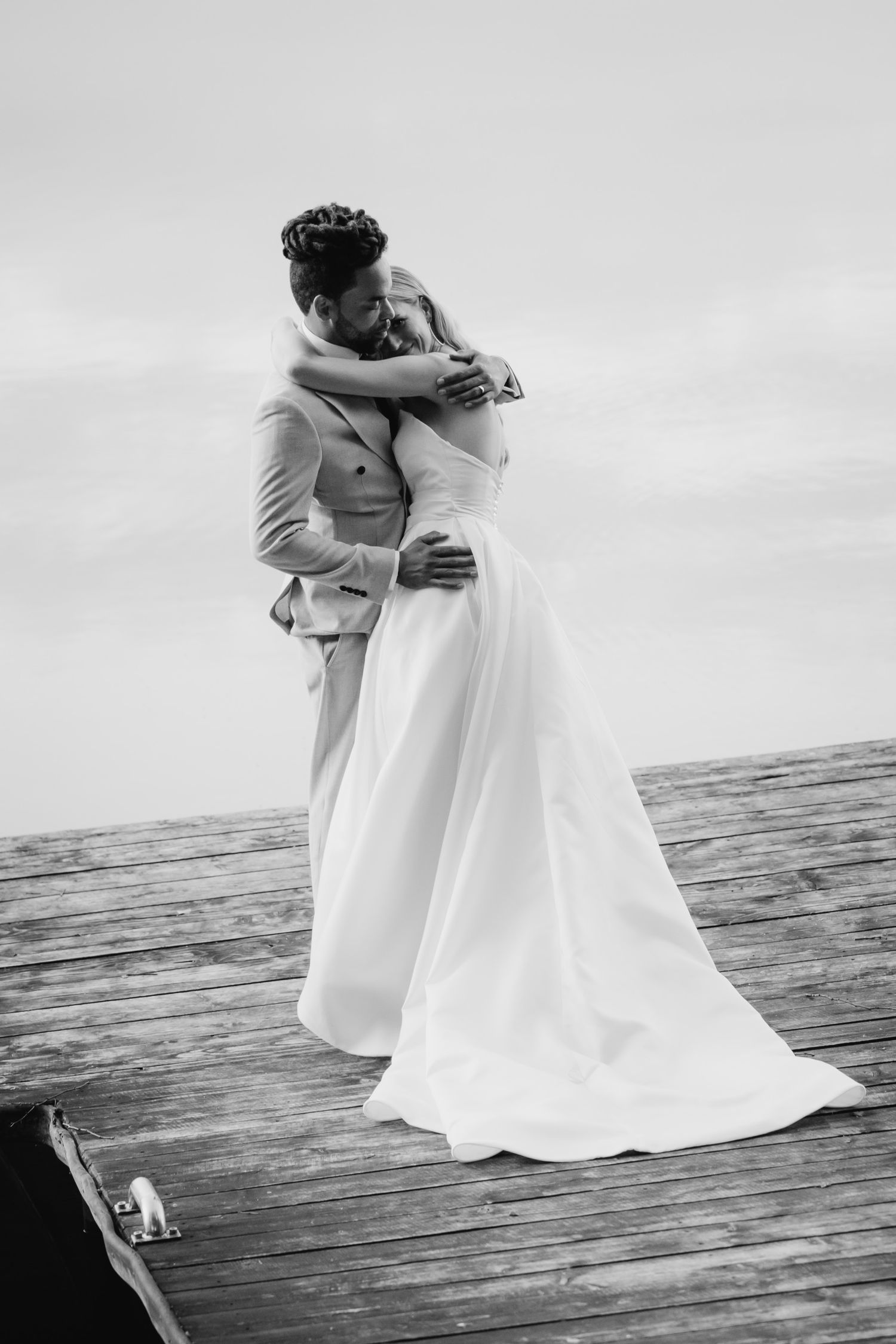 A tender black and white wedding moment captured on a wooden dock over water, with elegant white dress flowing in the breeze.