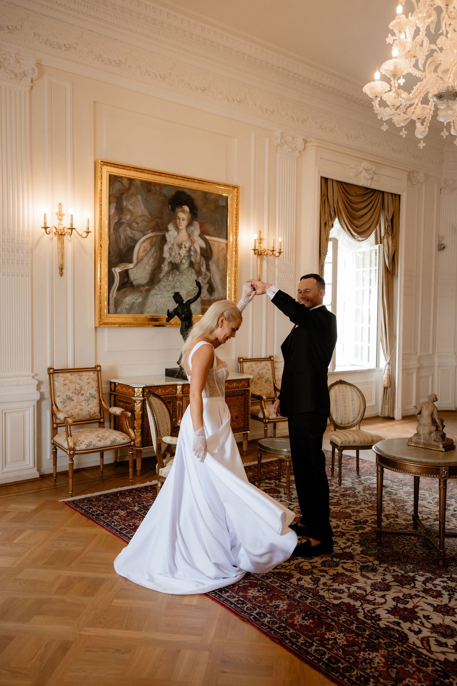 Couple dancing in an elegant room with ornate wall decor and chandelier.