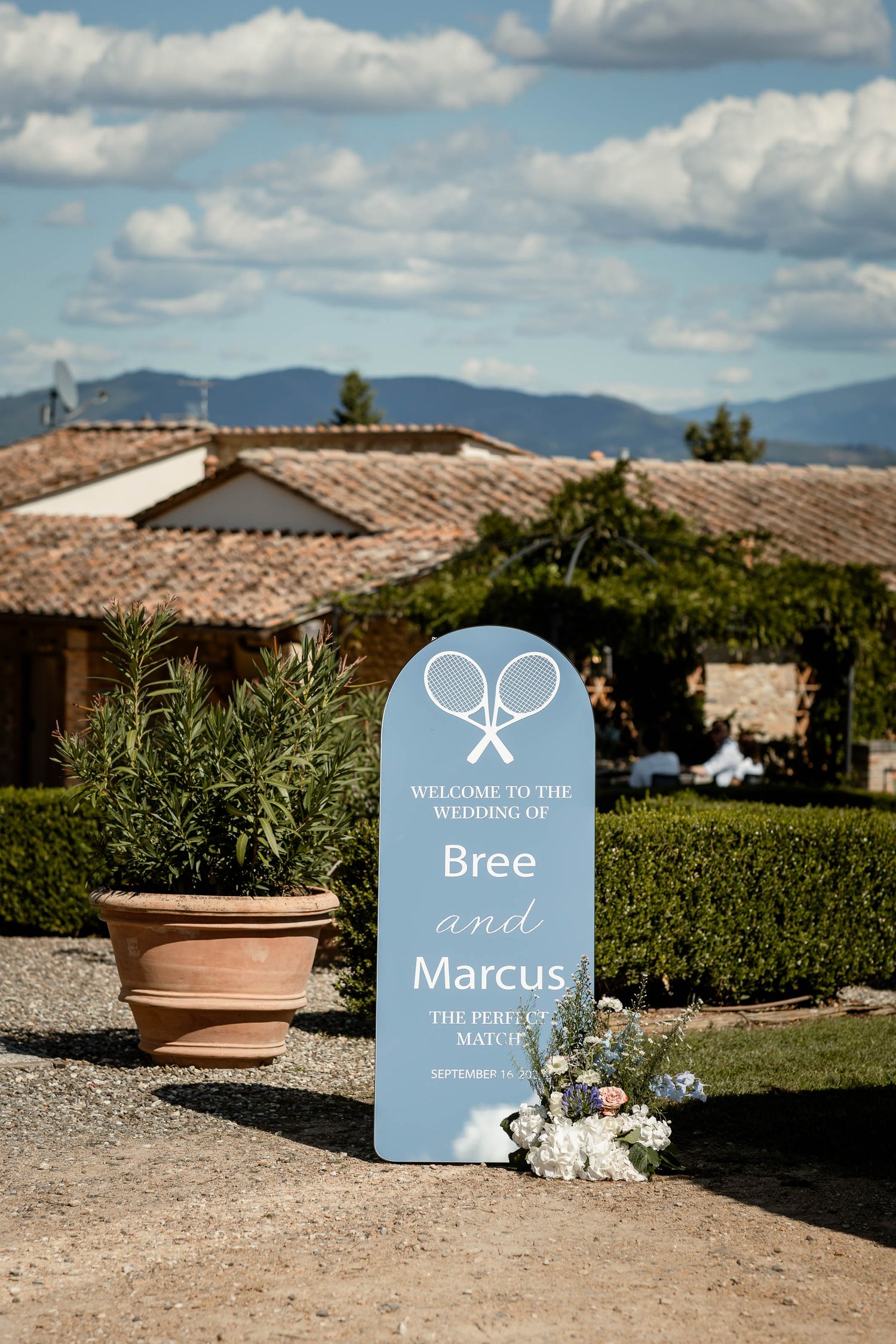 Blue wedding welcome sign with tennis racquet design against rustic Tuscan villa backdrop.