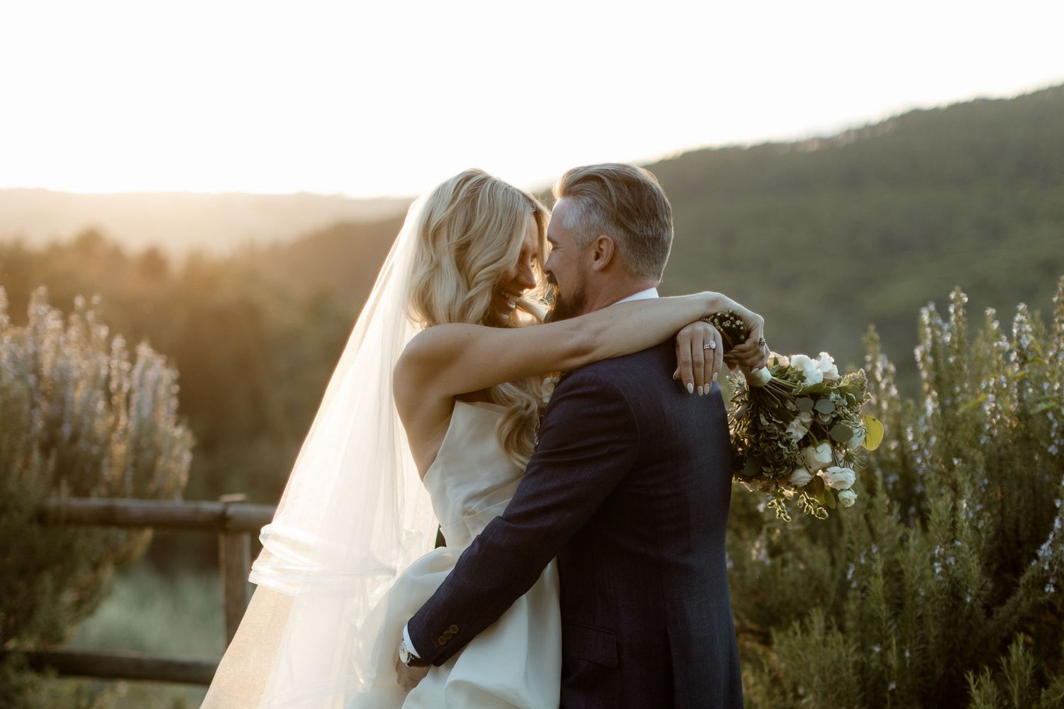 Couple in wedding attire embraces during golden hour sunset in a scenic outdoor setting.