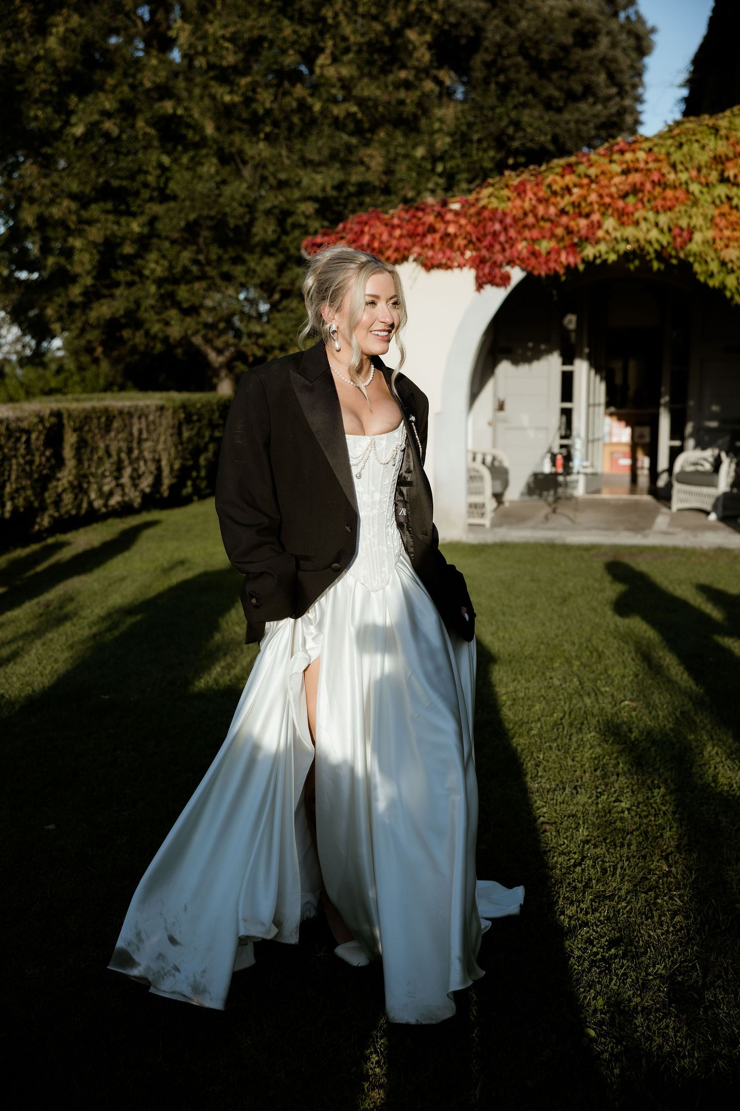 A couple in wedding attire dances and twirls in a sunlit garden setting with autumn foliage in the background.