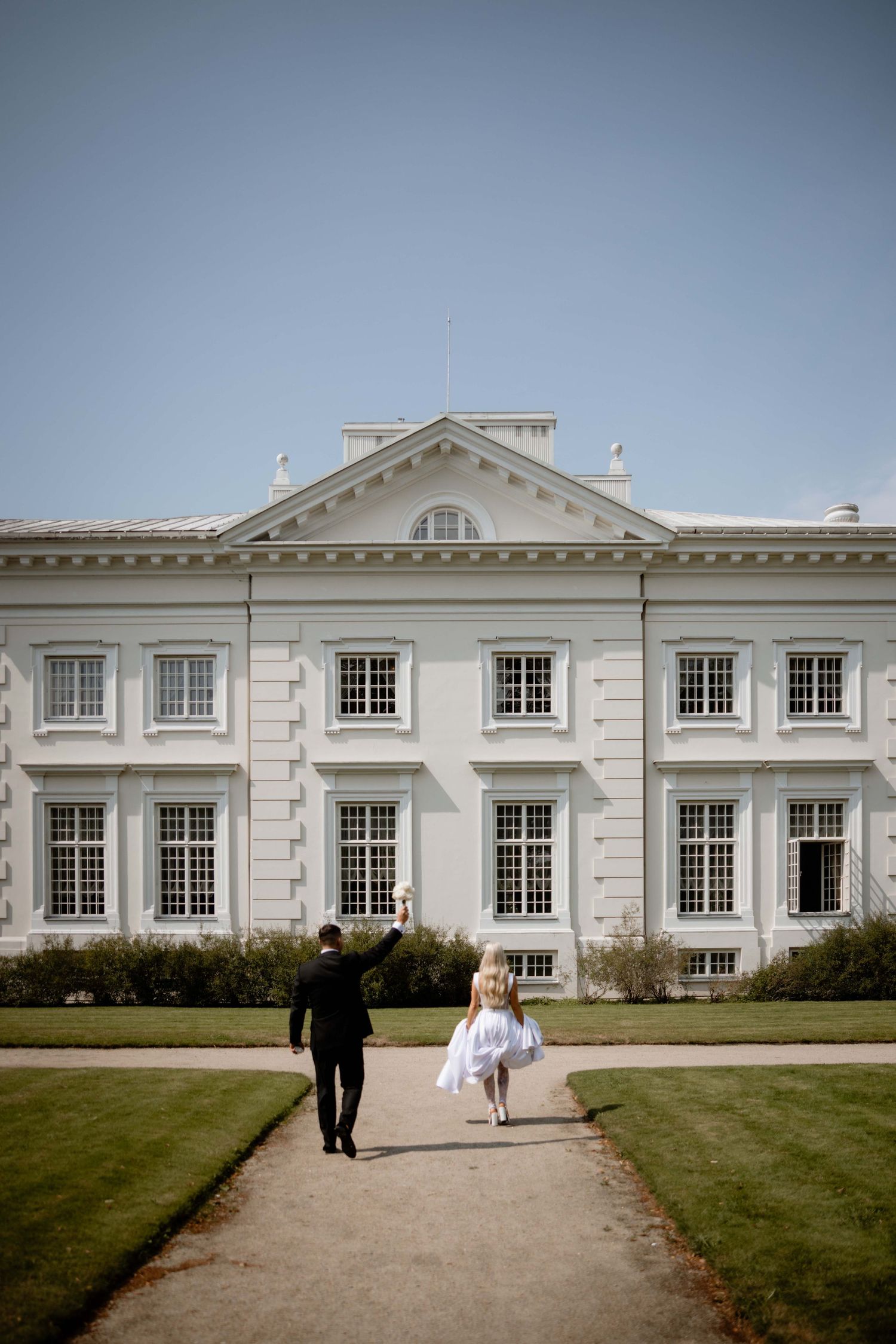 Two people walk along a path leading to an elegant white Georgian-style mansion with symmetrical windows and architecture.