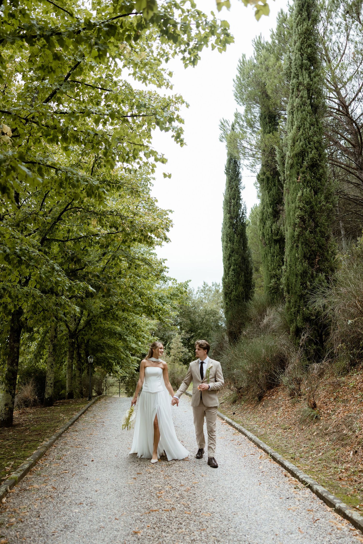A couple walks hand in hand down a cypress-lined gravel path in a romantic garden setting.