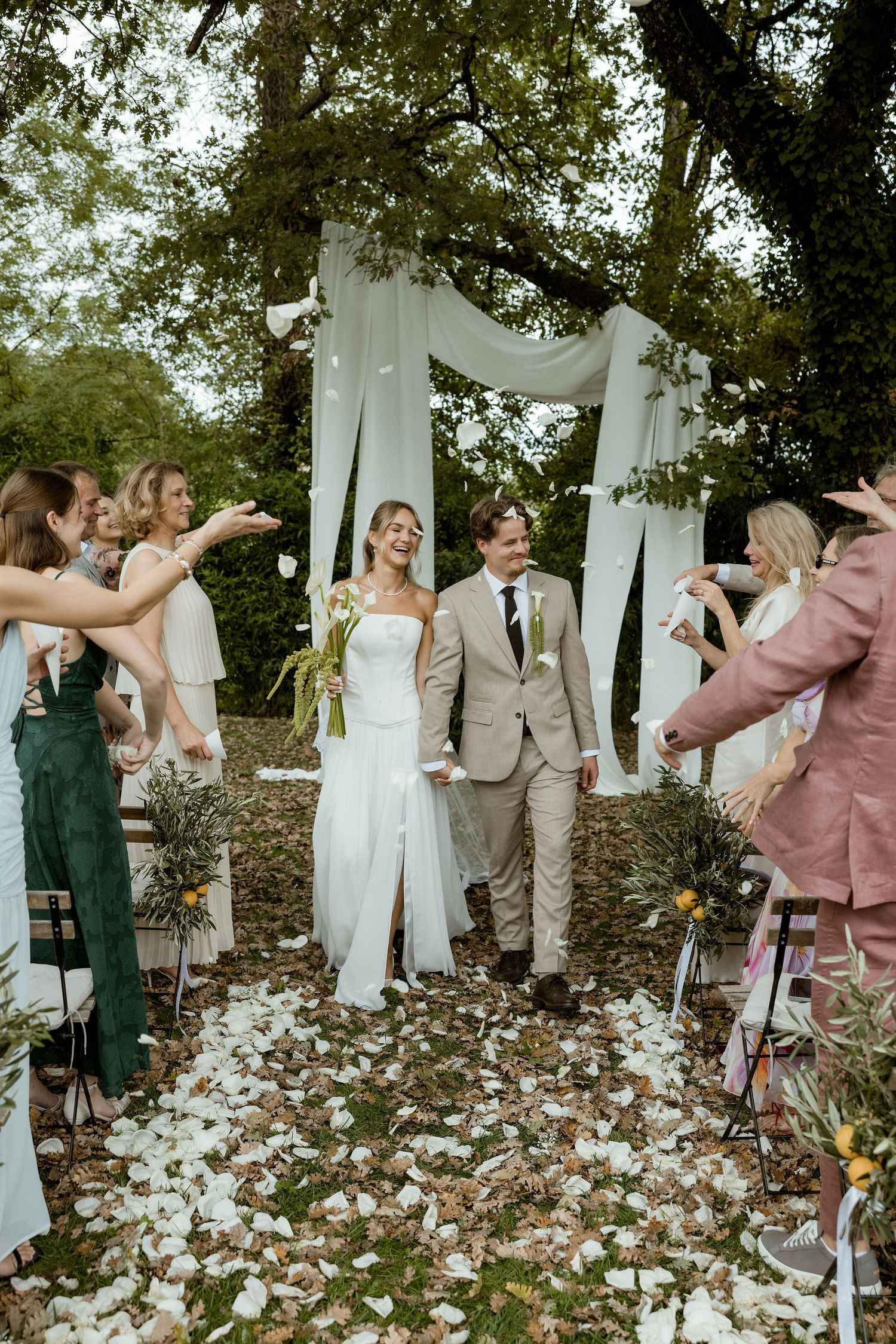Wedding party celebrates outdoors with white drapes and scattered flower petals creating a romantic garden atmosphere.