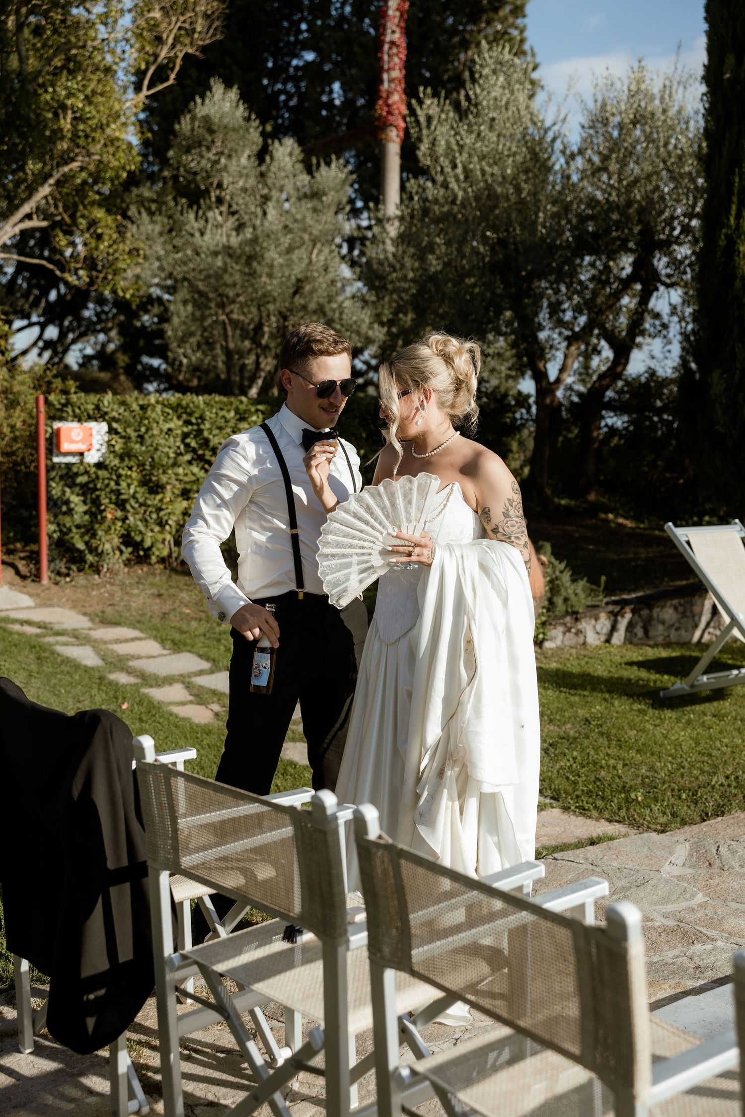 A couple in formal attire stands outdoors at a garden wedding venue on a sunny day.