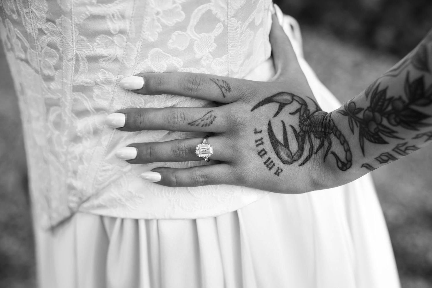 Black and white photo showing hands with wedding rings and tattoos against a white dress background.