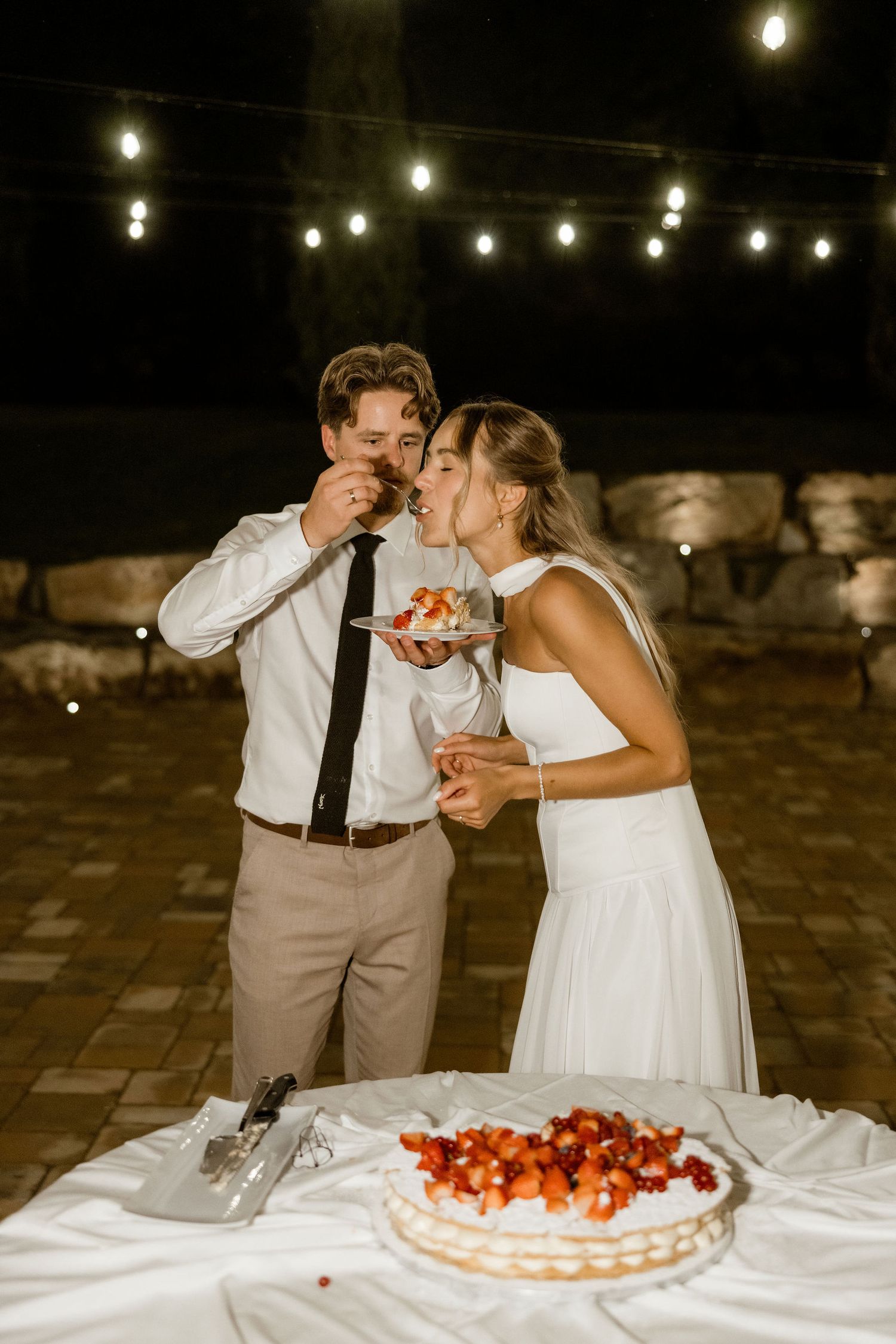 A couple in wedding attire playfully feed each other bites of strawberry cream cake at an outdoor evening reception.