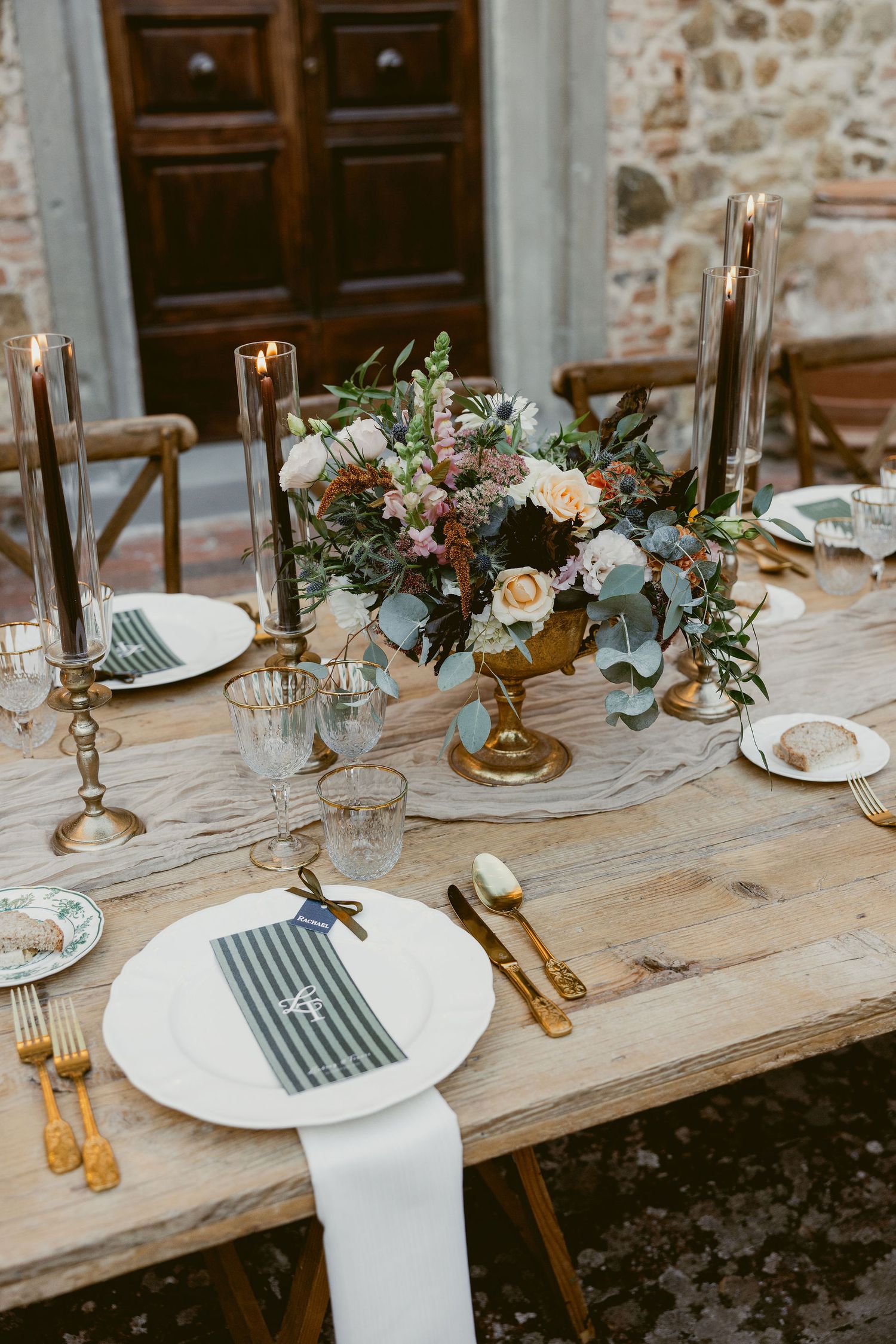 Rustic wedding table setting with white plates, gold candlesticks and delicate floral arrangements on a wooden farmhouse table.