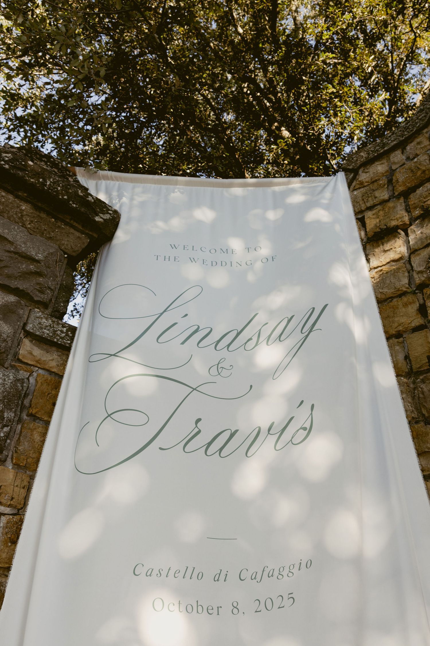 White wedding banner with elegant calligraphy text displayed against stone wall and trees in outdoor setting.