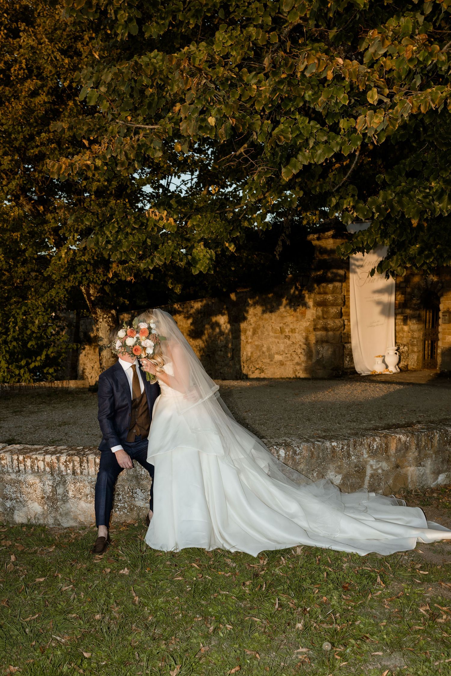 A couple in wedding attire poses together on a stone wall during an autumn sunset with trees in the background.