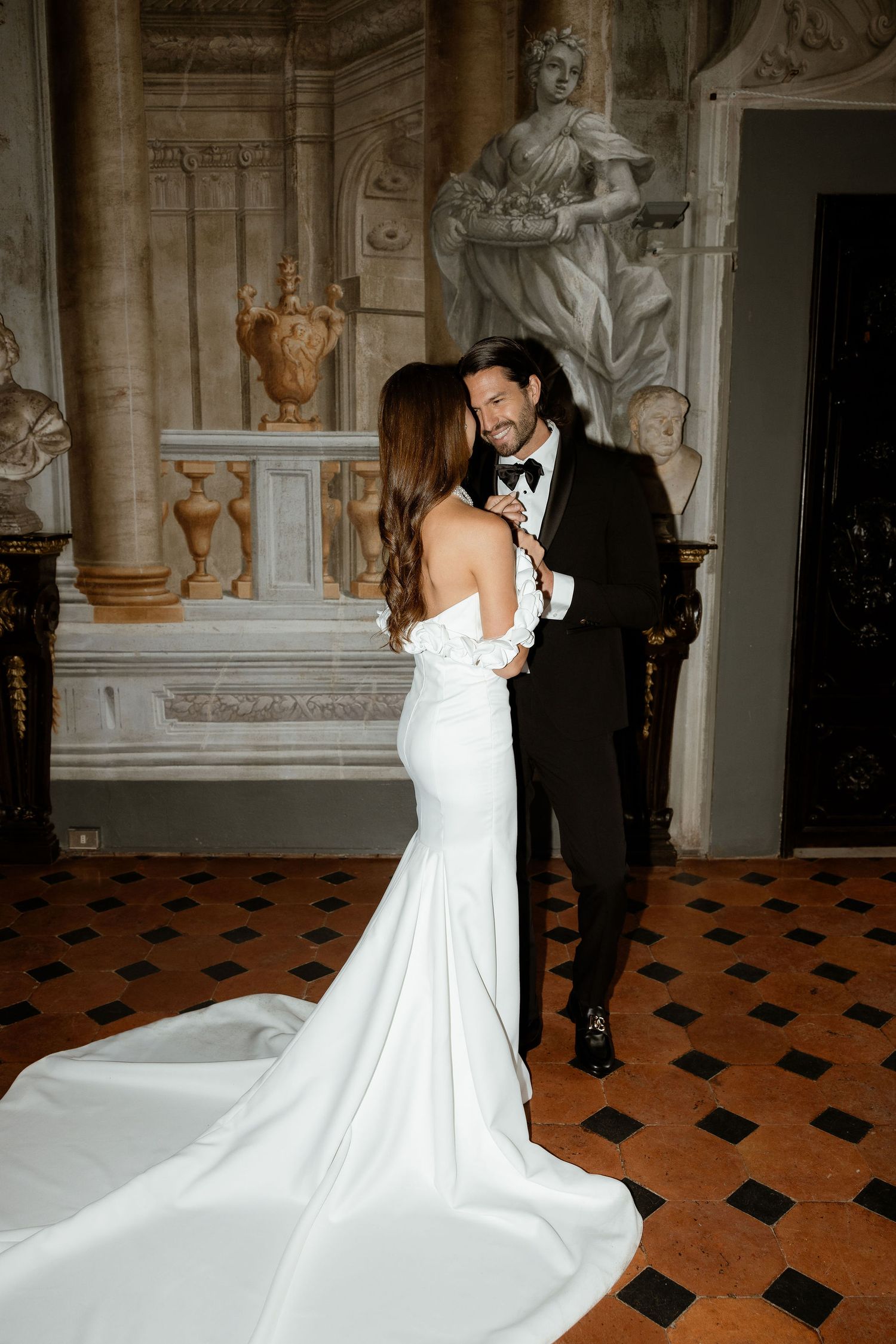 Elegant couple in formal wedding attire share intimate moment in ornate marble hall with classical statues and tiled floor.