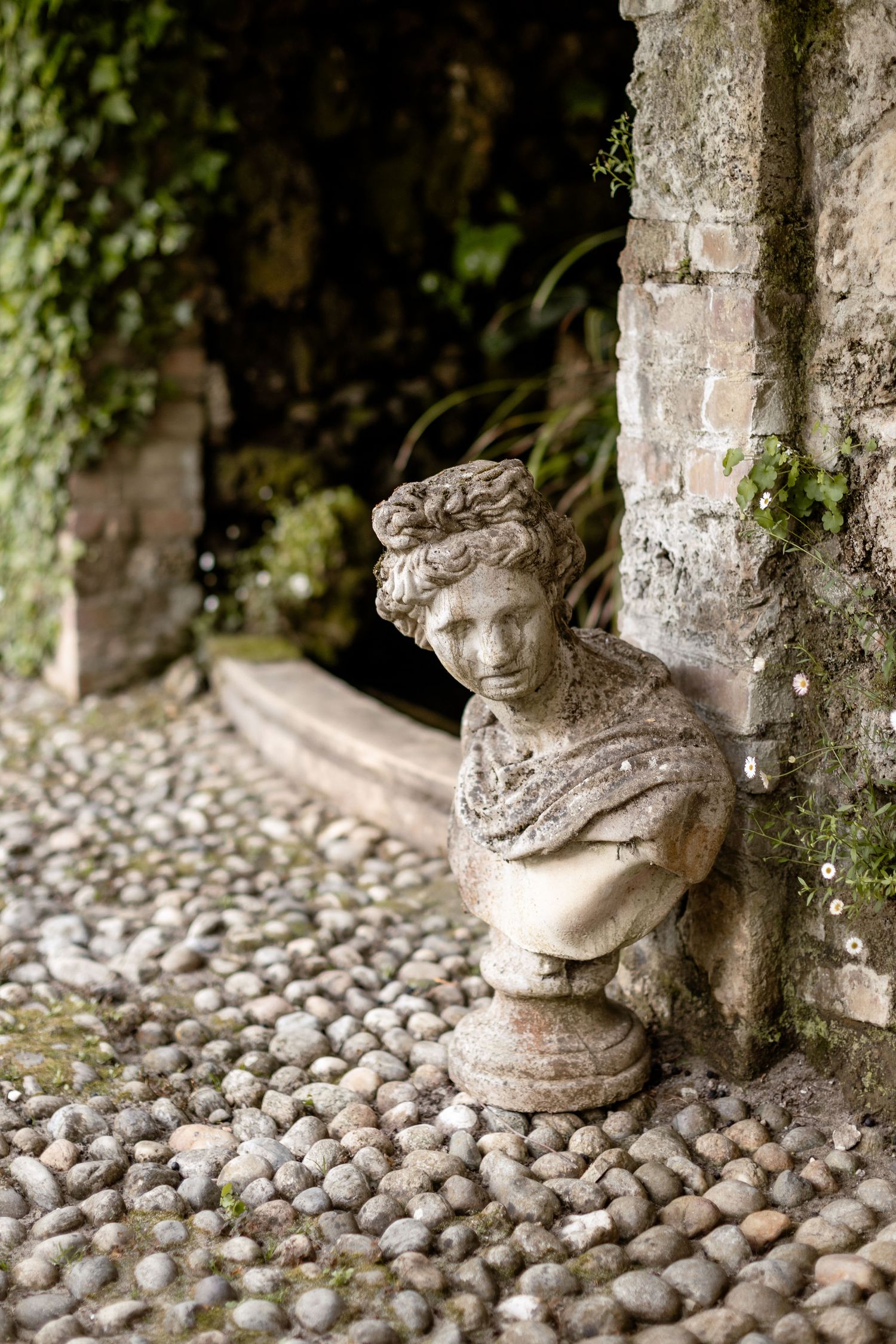 Classical stone bust sculpture on cobblestone path with ivy-covered wall in an ornate garden setting.