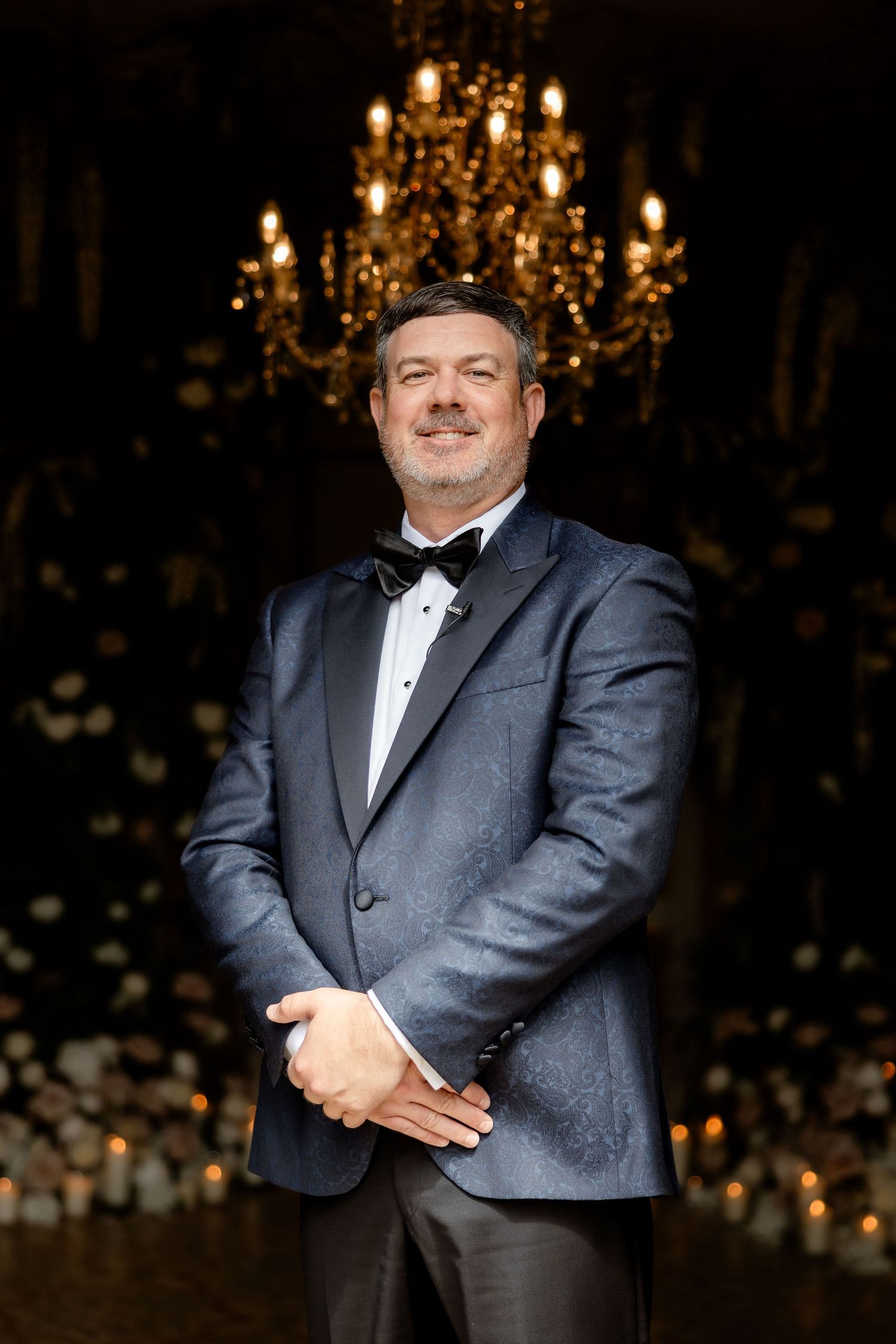 A person in a formal black tuxedo and bow tie stands in front of an elegant chandelier in a dark room.